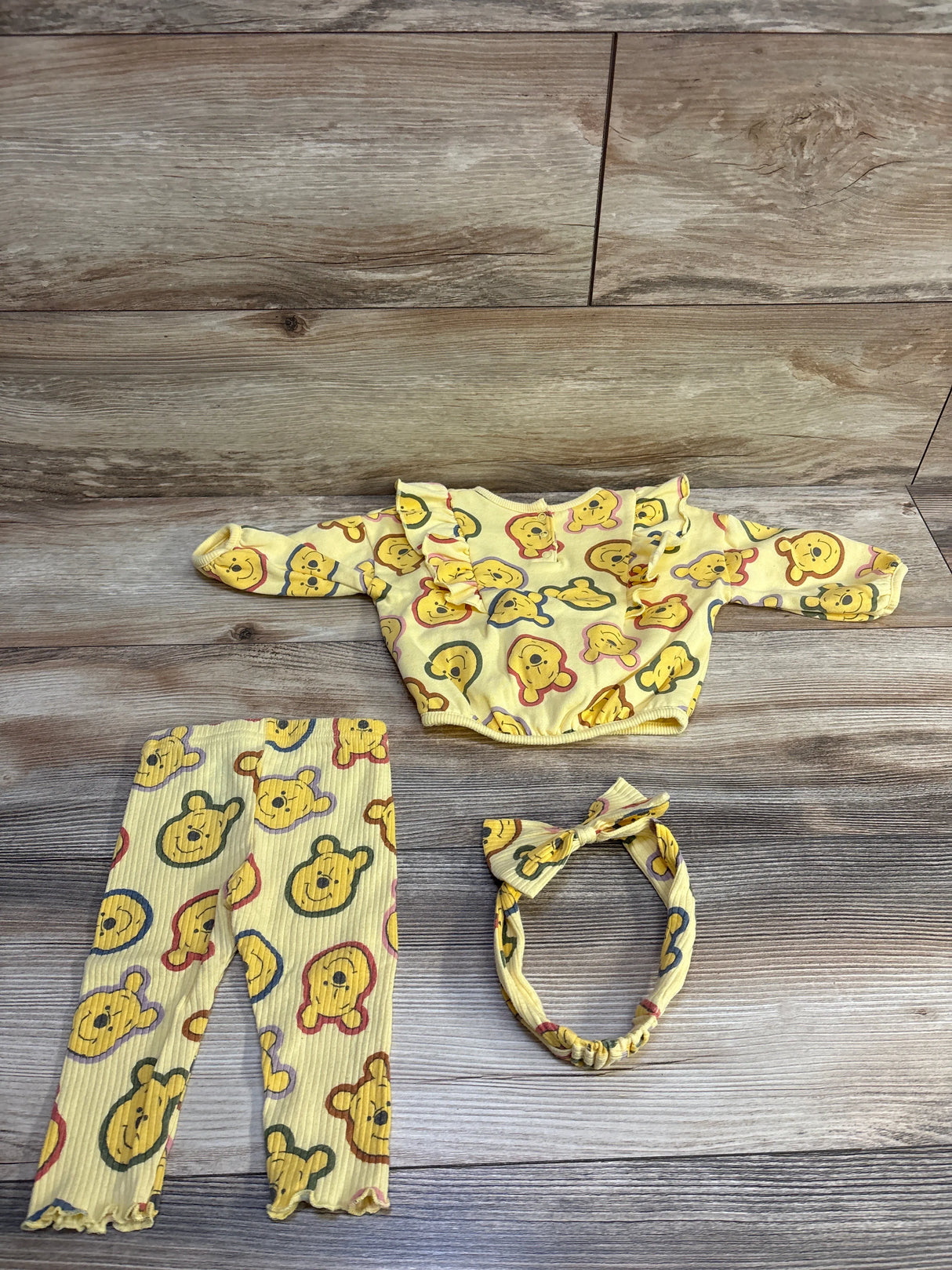 Flat-lay of yellow 3-piece Winnie the Pooh baby set: ruffled sweatshirt, ribbed leggings, and knotted headband on wood background