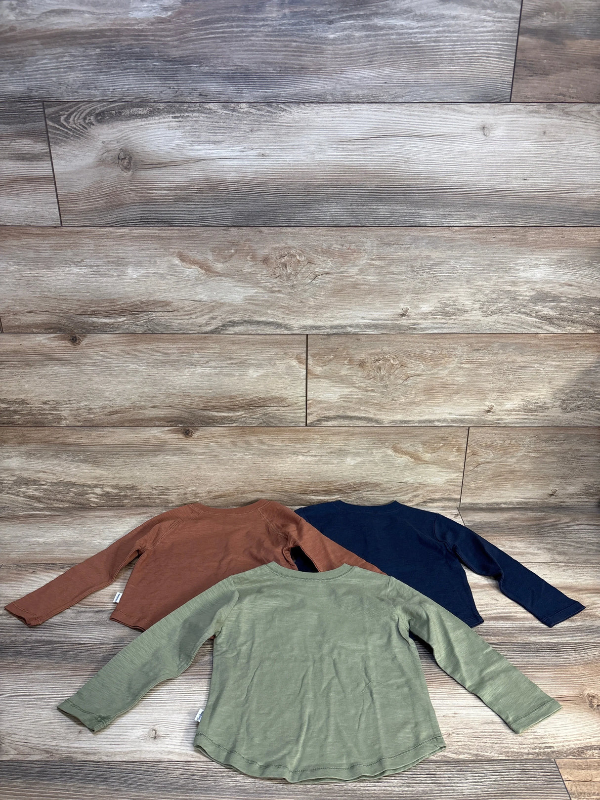 Back view of three Gerber long-sleeve Henley knit shirts (3T) in olive green, rust brown, and navy on wood floor