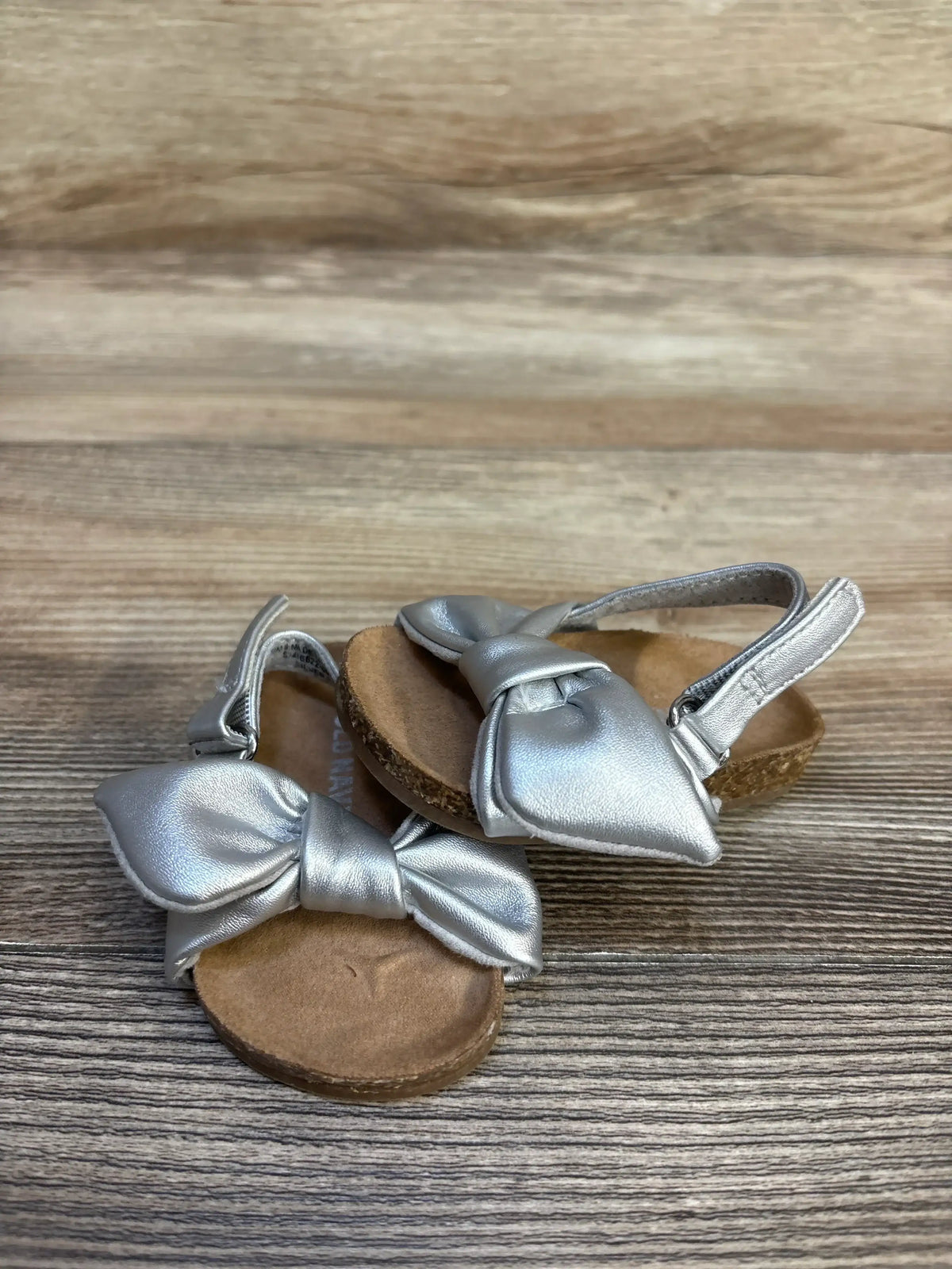 Angled view of Old Navy faux-leather tie-bow toddler sandals in silver highlighting bow detail and cork soles on wood background