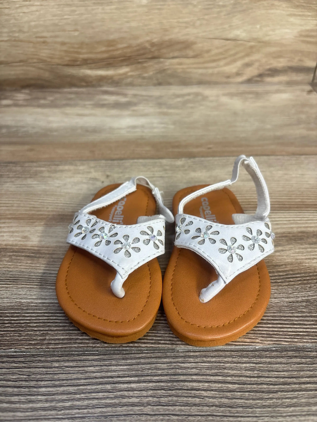 Front view of Capelli white backstrap sandals showcasing floral embellishments and brown soles on a wood surface