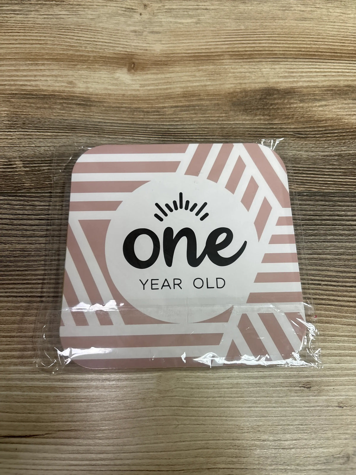 Front view of a 12pc monthly milestone photo prop card showing one year old text and pink geometric border on wood