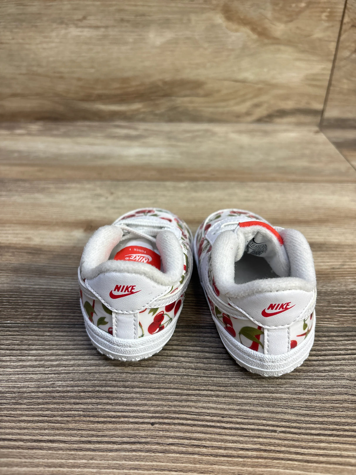Rear view of white Nike baby crib shoes with red cherry print, showing red Nike heel logos and padded collars on wood floor