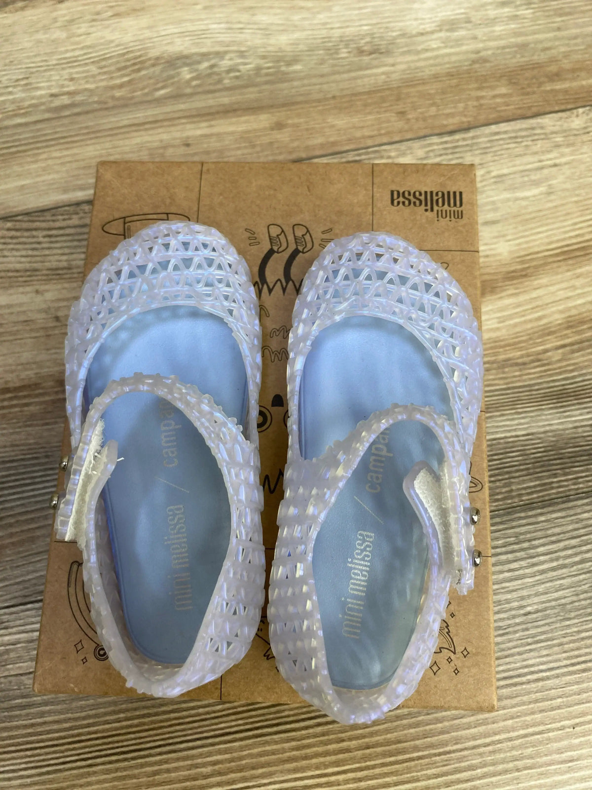Top-down view of Mini Melissa Campana Papel jelly flats in pearly blue showing lattice cutout upper and Mary Jane strap on cardboard box
