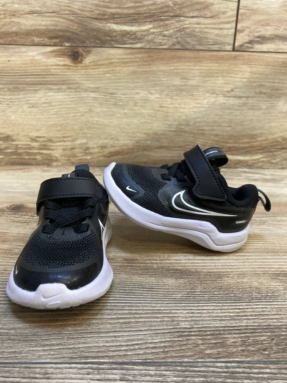 Side view of Nike Cosmic Runner TD toddler sneakers in black and white showcasing swoosh logo velcro strap and white sole on wood plank floor