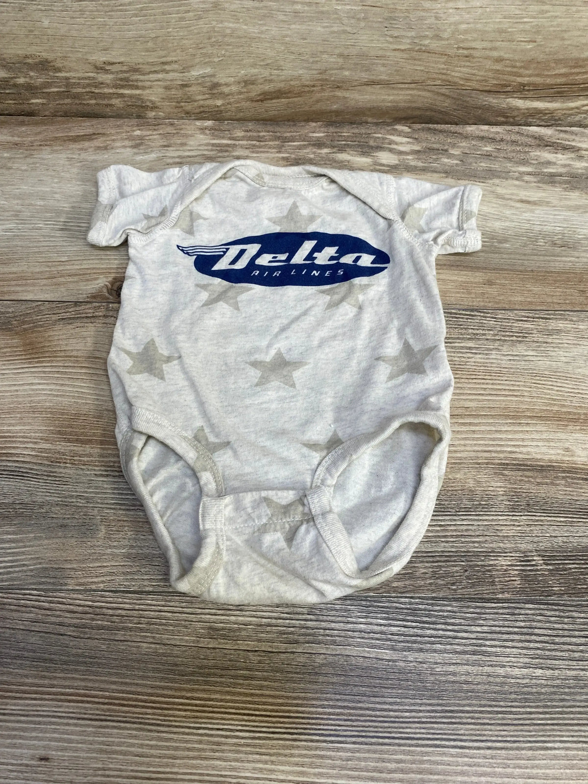 Flat lay of oatmeal short-sleeve baby bodysuit (3-6M) showing blue Delta Airlines logo and subtle star print on wood floor