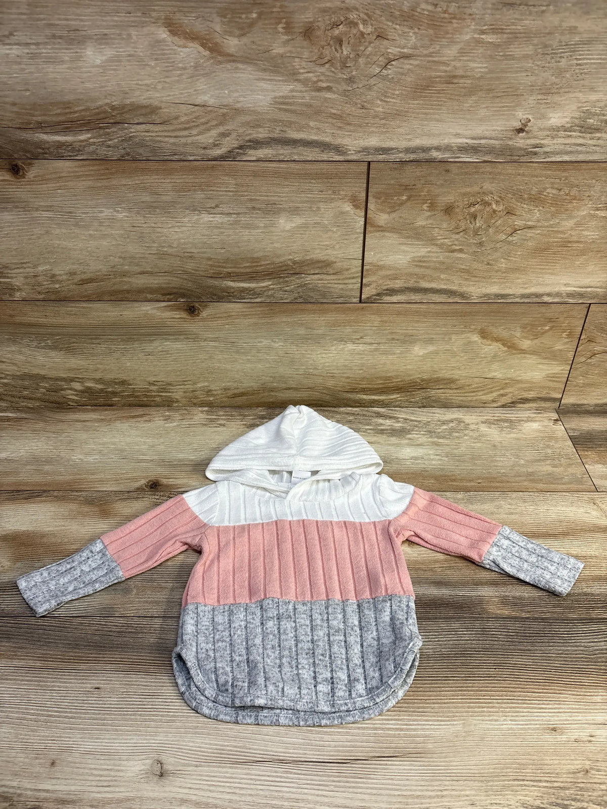 Flat lay of toddler hooded sweater showcasing ribbed knit and colorblock panels with white hood, pink middle, and gray hem and cuffs on wood background