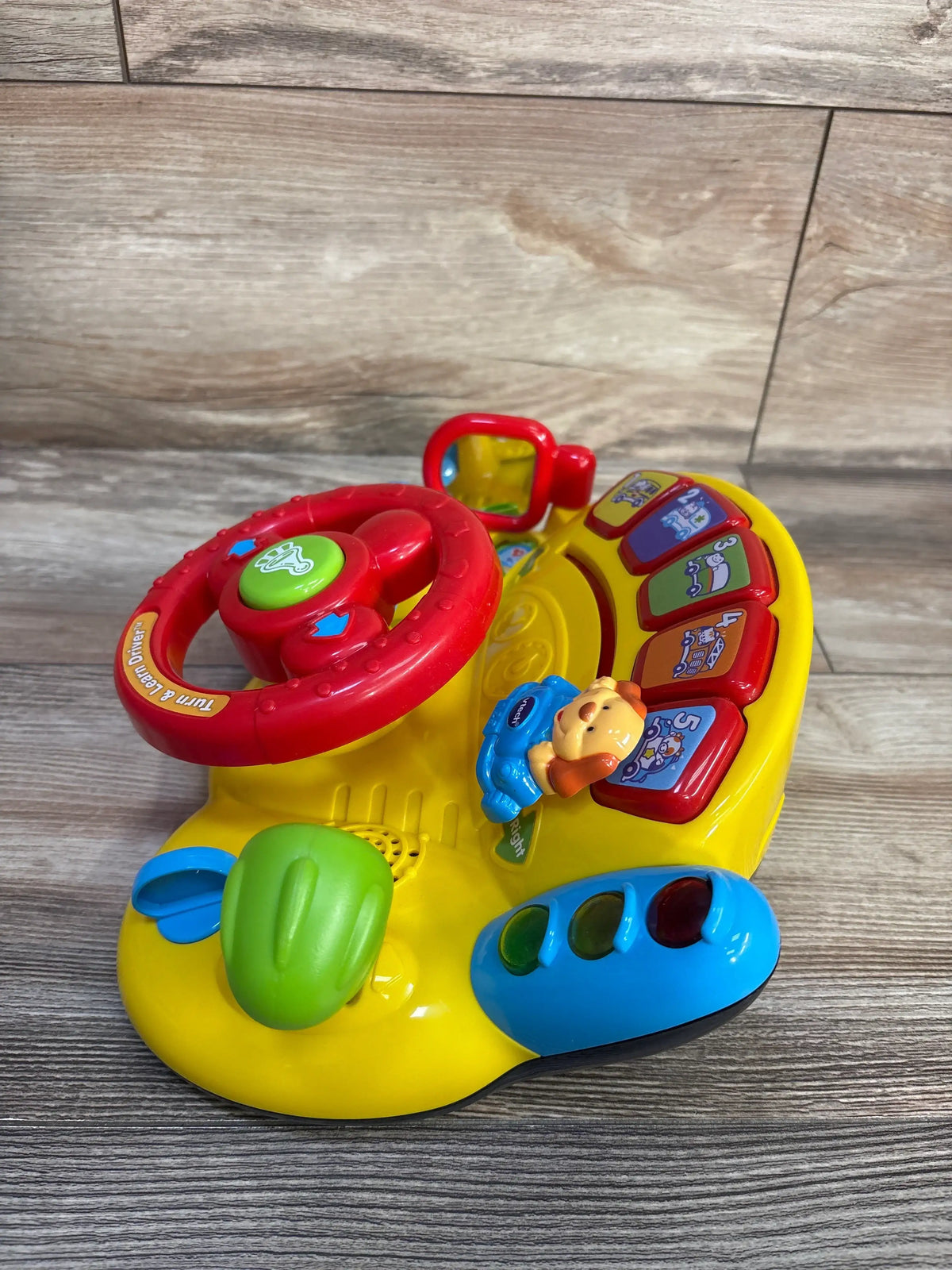 Three-quarter view of Vtech Turn and Learn Driver showcasing colorful controls, steering wheel, gear shift, and signal lever on a wood floor