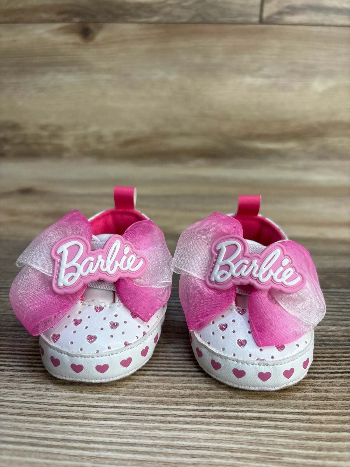 Front view of Barbie soft sole baby sneakers size 1C with pink organza bows, Barbie logo, and pink heart print on white upper, wood background