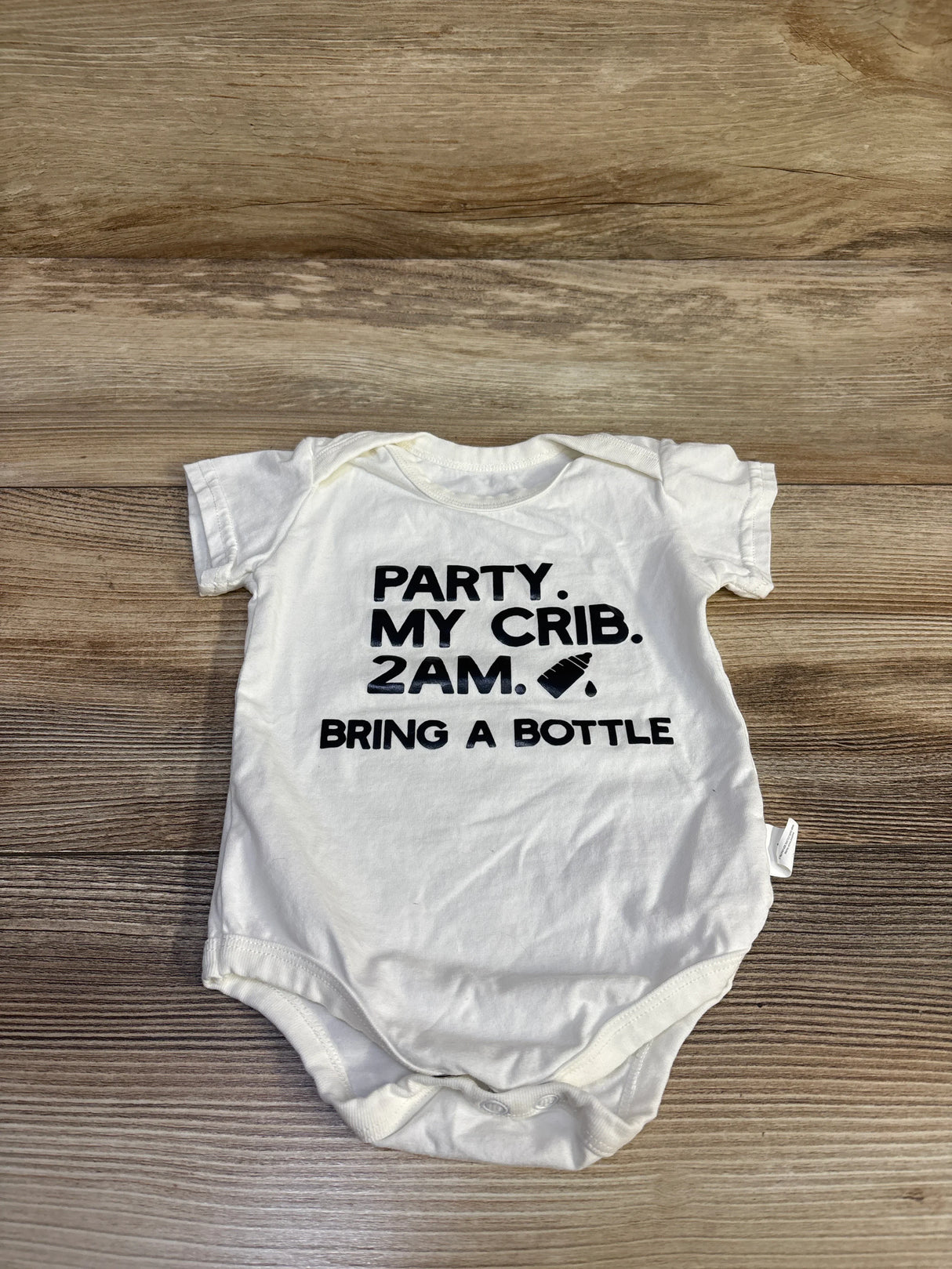 Flat lay of white baby bodysuit on wood background showing black slogan text: PARTY. MY CRIB. 2AM. BRING A BOTTLE