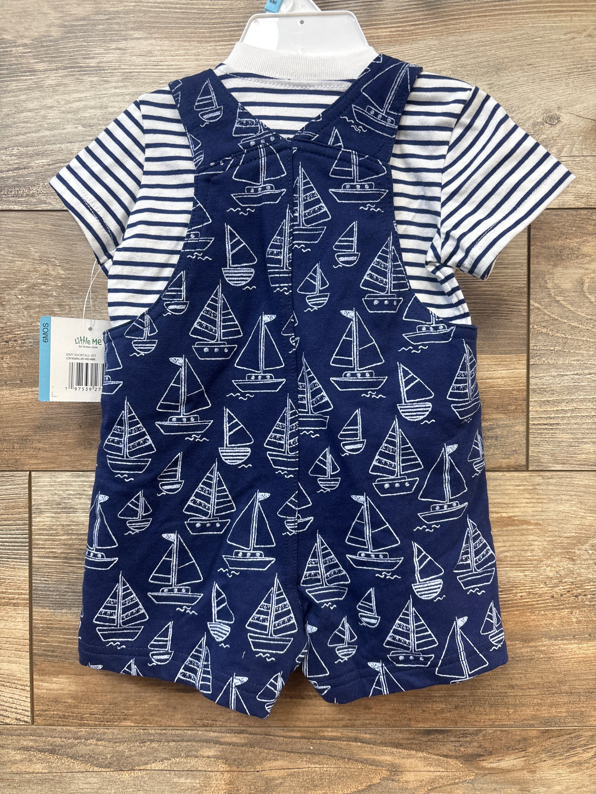Front view of Little Me two piece set with white and navy striped shirt and navy sailboat print shortalls on wood background