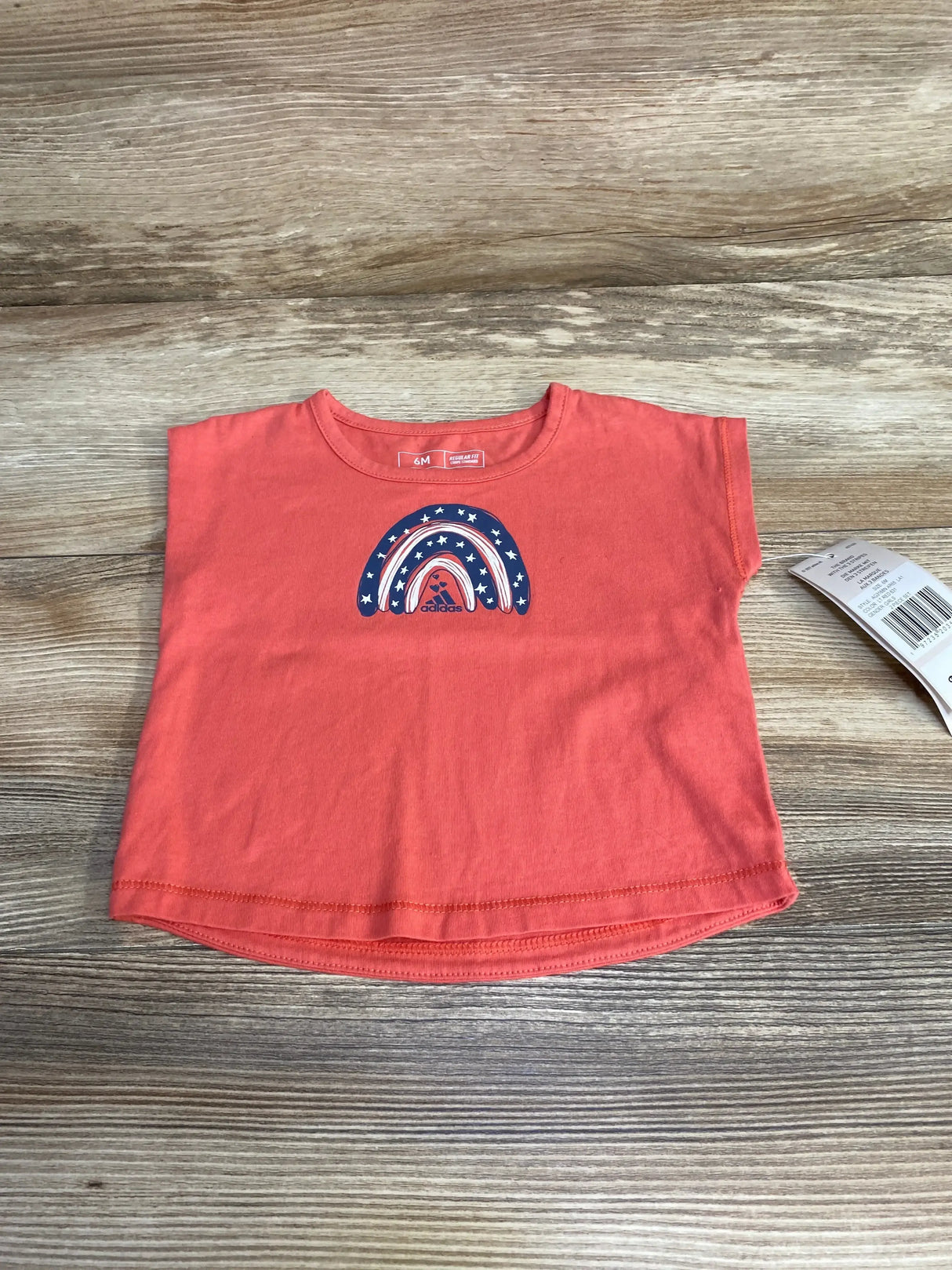 Flat lay of red Adidas baby tee showing rainbow graphic and short sleeves on wood plank background with size tag visible