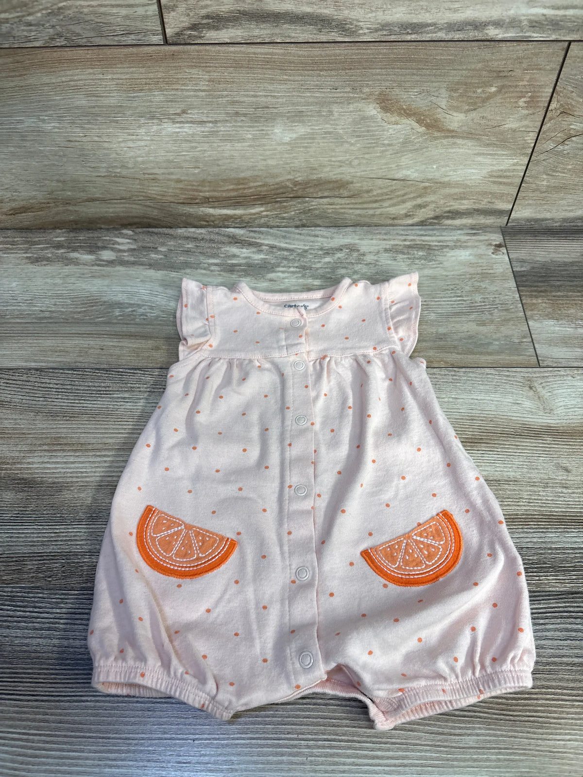 Front view of Carter’s button-front shortie romper showcasing peach fabric, orange dots, and citrus slice pockets on a wood floor