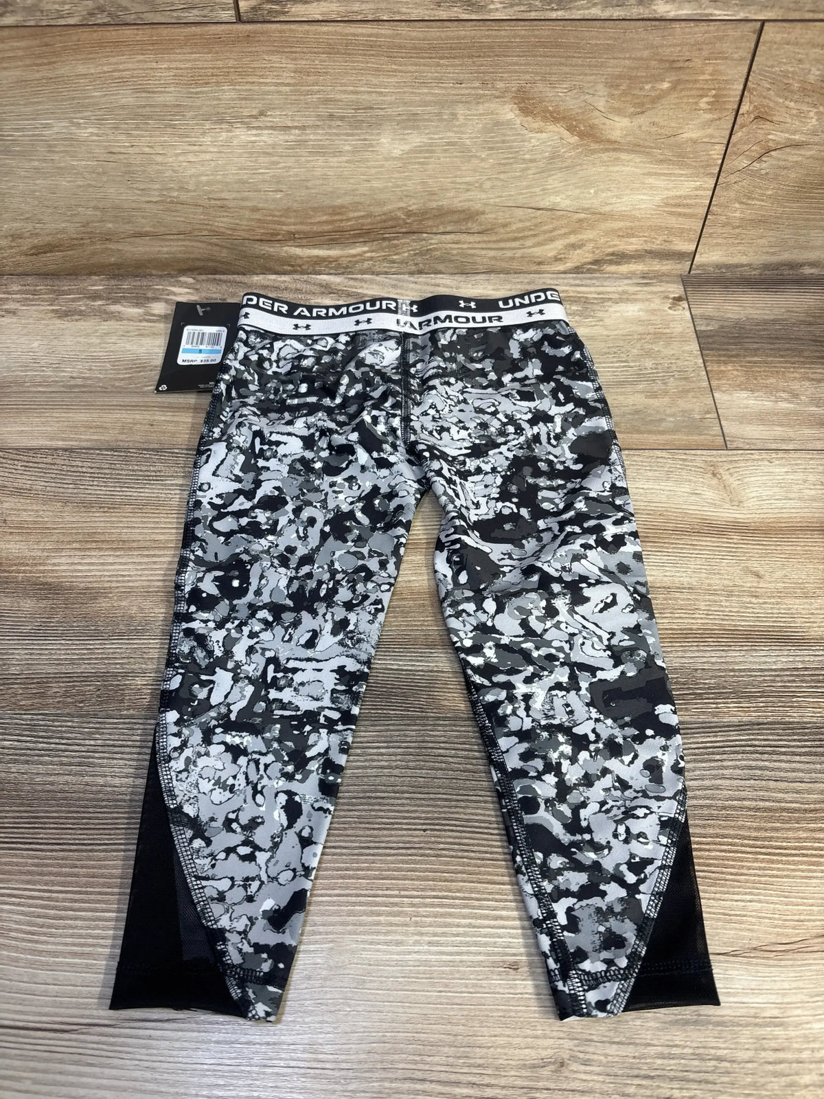 Flat lay of Under Armour girls' grey camo leggings (size 5T) showing branded elastic waistband and black mesh ankle panels on wood floor