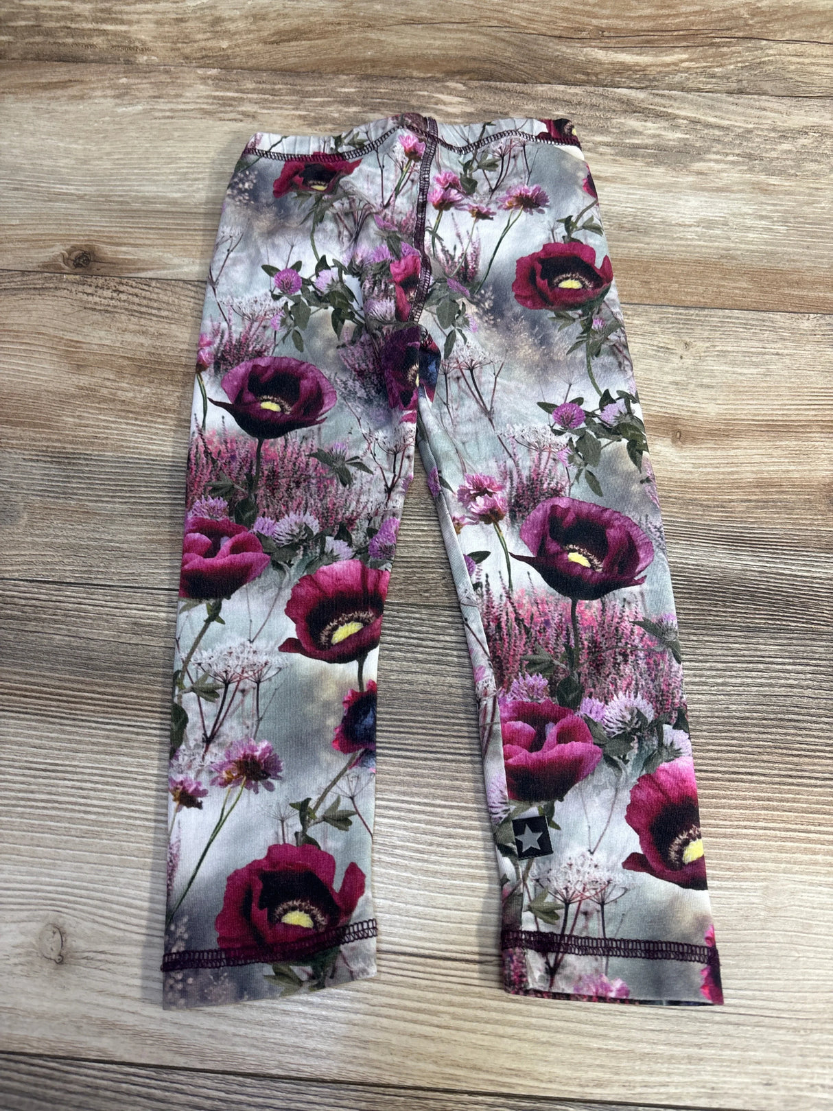 Flat lay of Molo Stefanie leggings size 2T showing pink and purple floral print, elastic waistband and ankle star logo on light wood floor