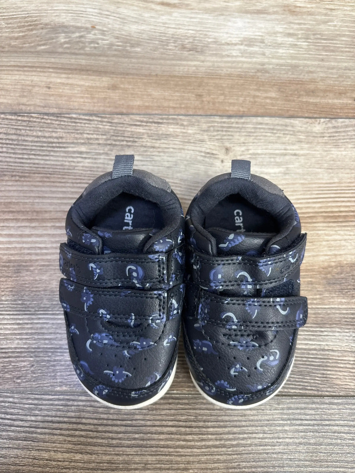 Top view of Carter’s Every Step First Walker Neo Dino sneakers with two hook and loop straps and pull tabs on wood floor