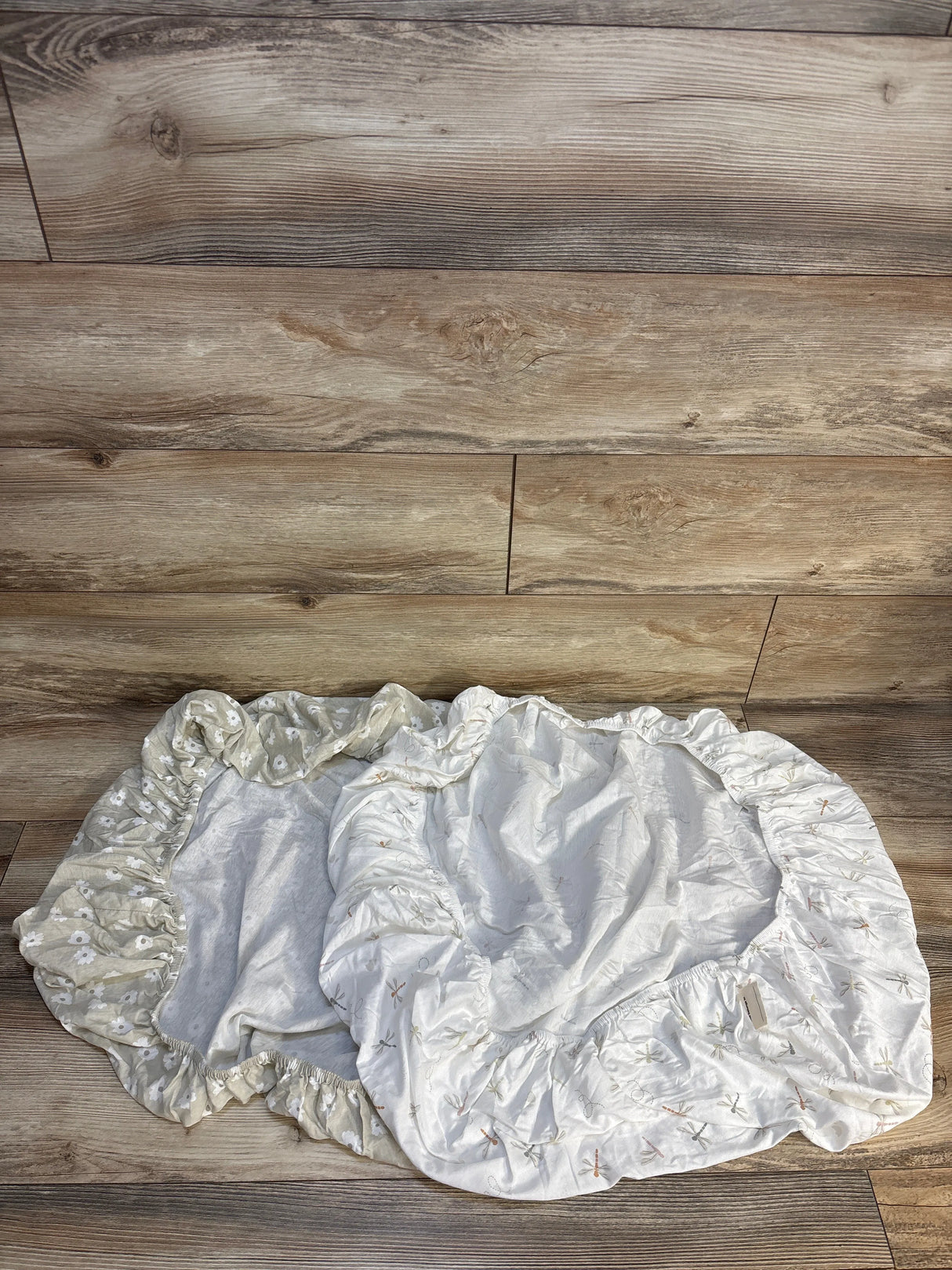 Top-down view of KeaBabies Soothe fitted mini crib sheets 2 pack in Meadow showing beige daisy print and white dragonfly print on wood floor