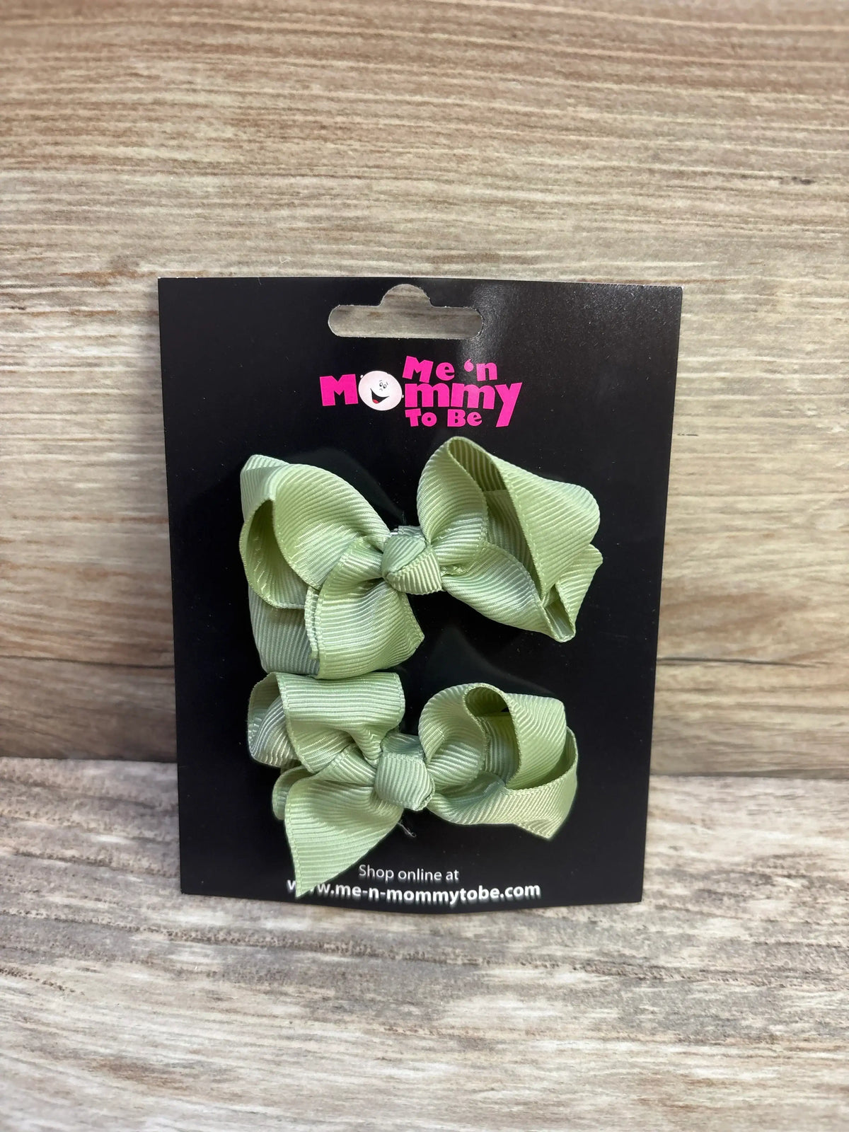 Front view of 2 light olive green grosgrain hair bows on black Me n Mommy To Be display card against wood background