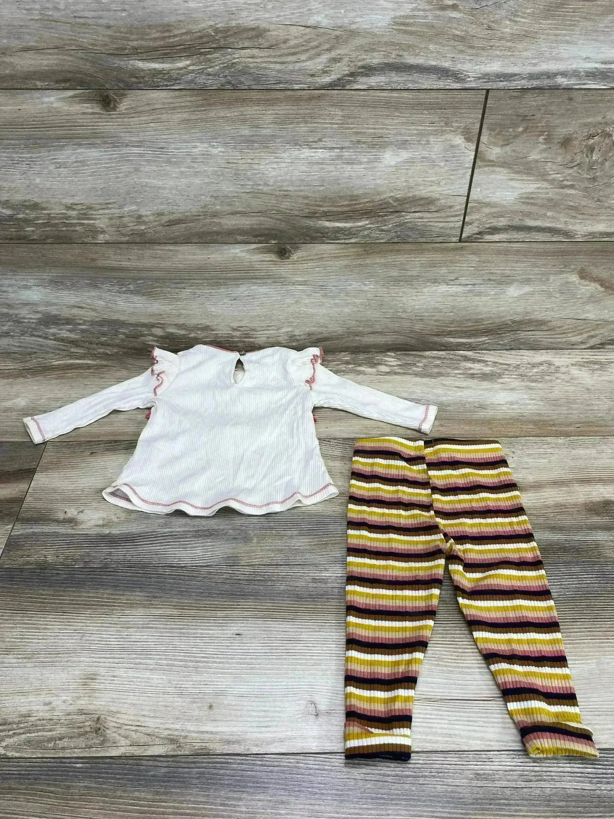 Flat lay of Mudpie 2-piece tassel top and striped ribbed leggings, cream top with ruffled sleeves and scalloped hem on gray wood floor