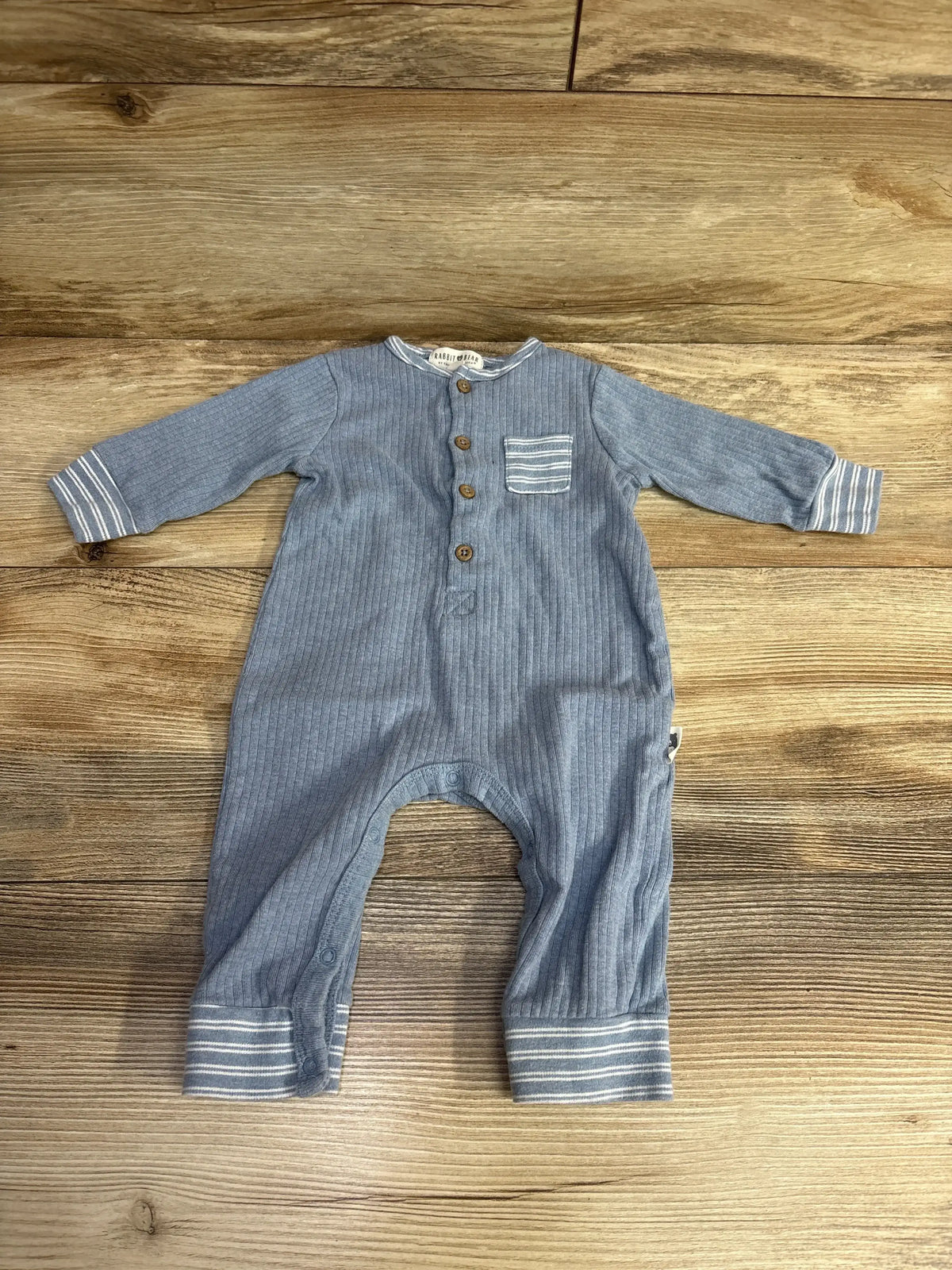 Blue ribbed baby romper with striped cuffs, wood buttons, and chest pocket on wood floor