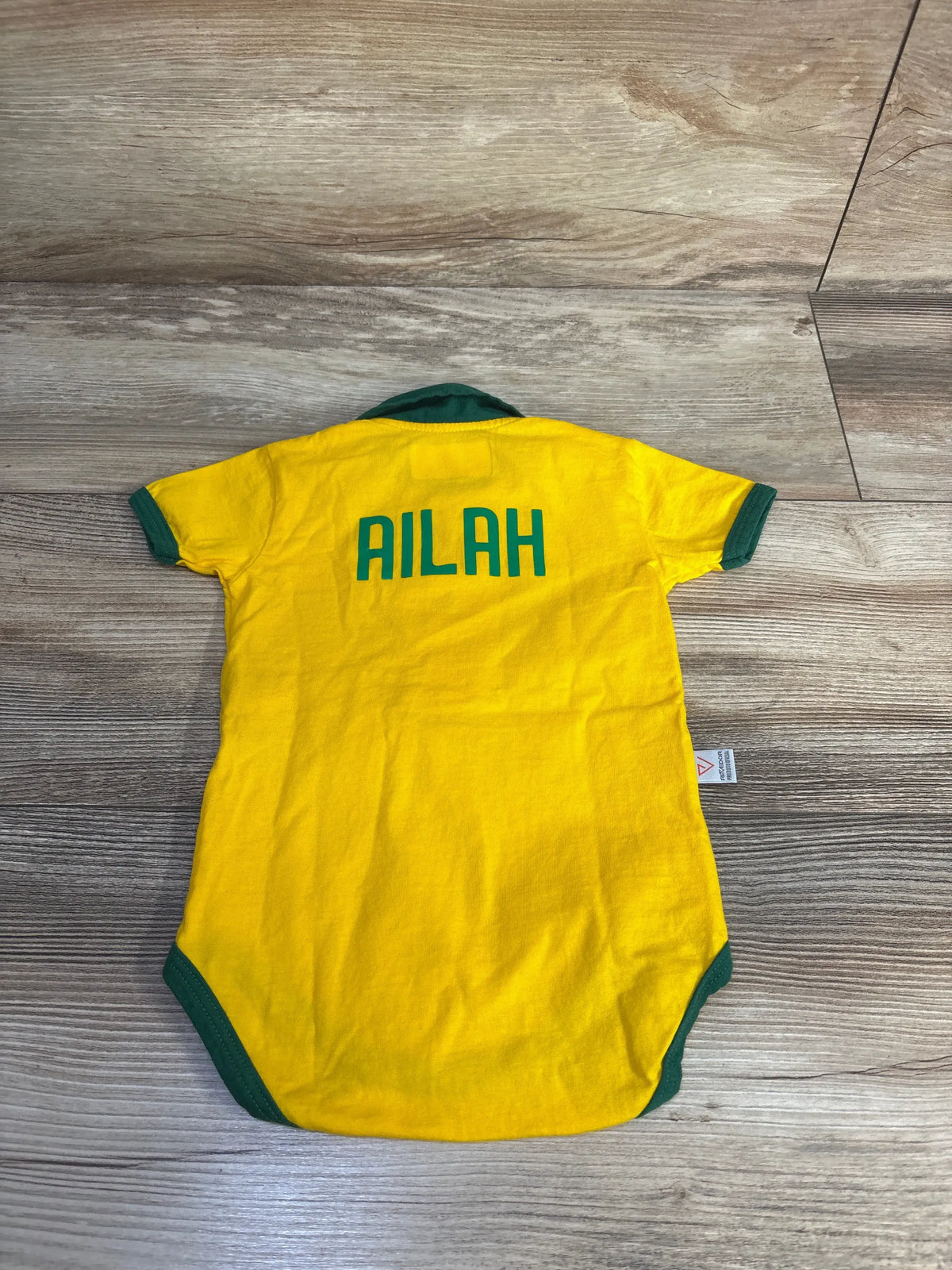 Back view of yellow Revedor Brazil polo bodysuit showcasing green trim and AILAH lettering on wood-grain background