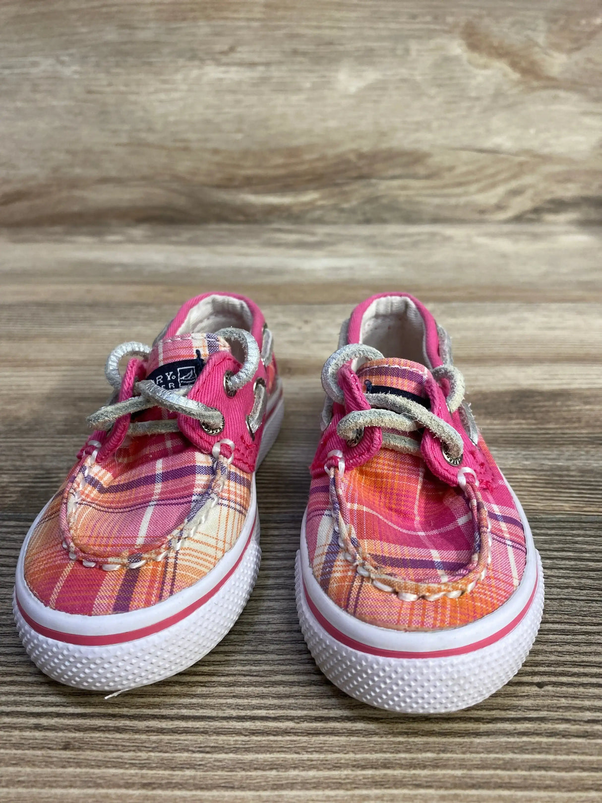 Front view of Sperry toddler boat shoes showing pink-orange plaid canvas white rubber soles, braided laces on wood background