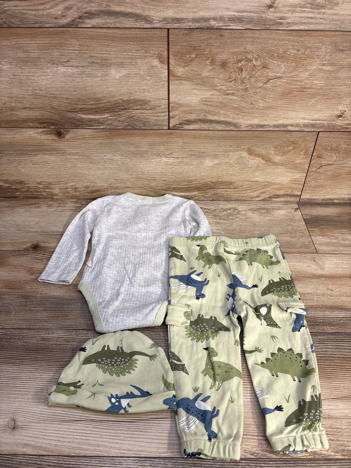 Top view of Kit + Pearl 3-piece grey ribbed henley set with dinosaur-print pants and matching hat on wood flooring