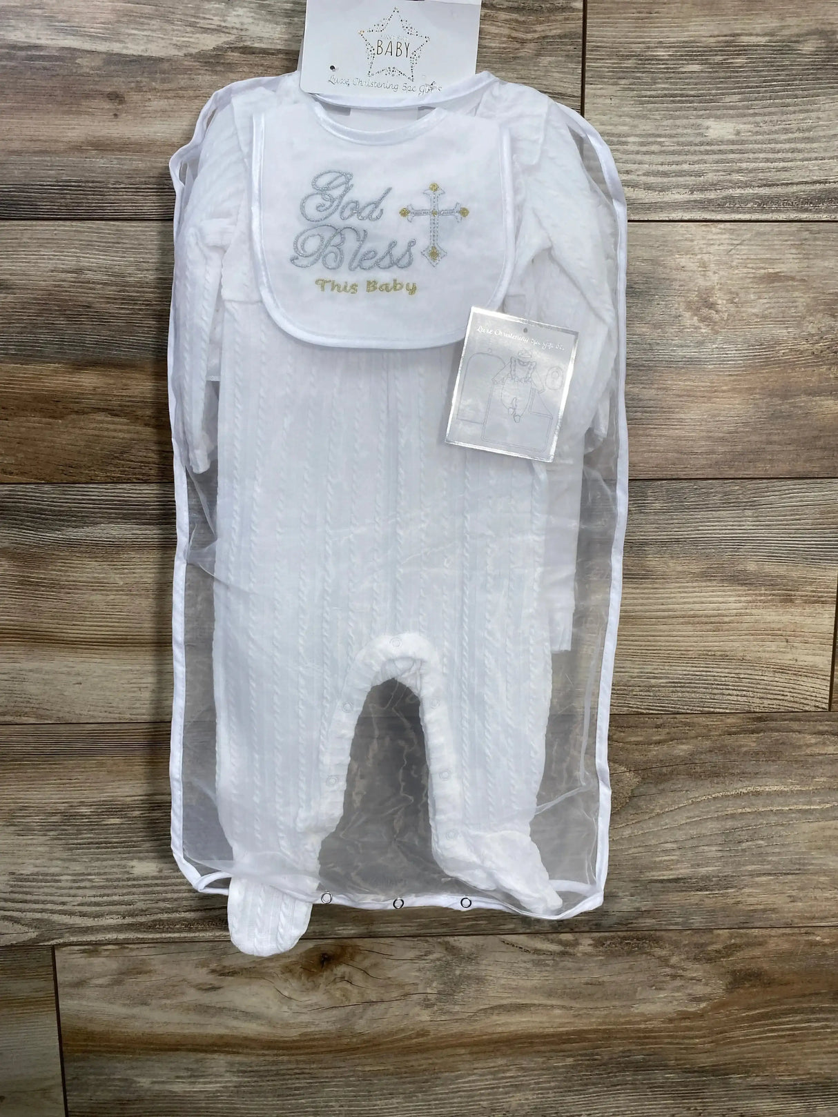 Flat lay of NEW Superstar Baby 5pc Luxe Christening Gift Set in white showcasing God Bless This Baby bib and footed sleeper on wood plank floor
