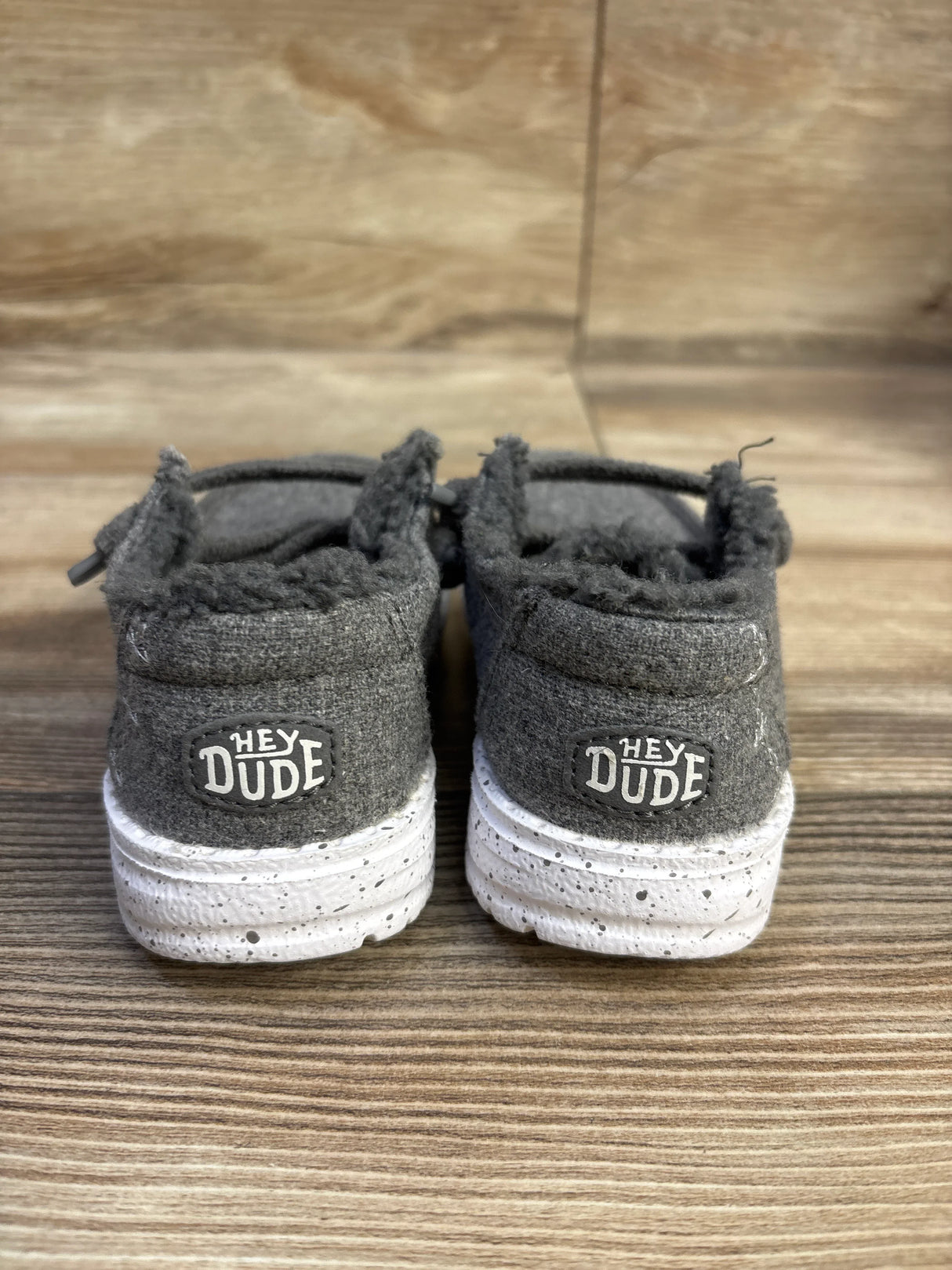 Rear view of Hey Dude Wendy kids wool slip-on size 7C showing fuzzy cuff, Hey Dude logo on heel, and white speckled sole on wood background