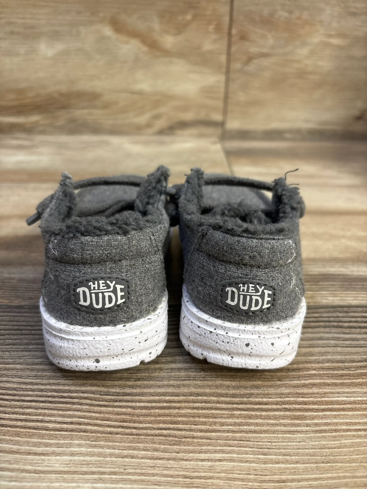 Rear view of Hey Dude Wendy kids wool slip-on size 7C showing fuzzy cuff, Hey Dude logo on heel, and white speckled sole on wood background