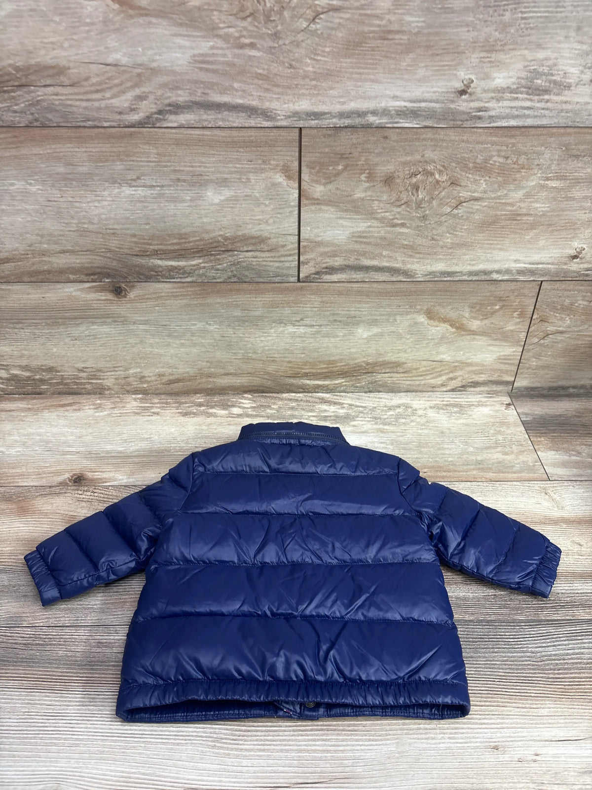 Back view of a navy blue puffer jacket for babies, size 9m, on a wooden background.