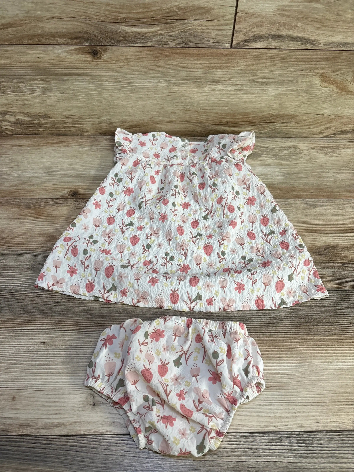 Overhead view of Deux Par Deux baby dress and bloomers set showcasing an allover flower and strawberry print with puffed sleeves on a wood background