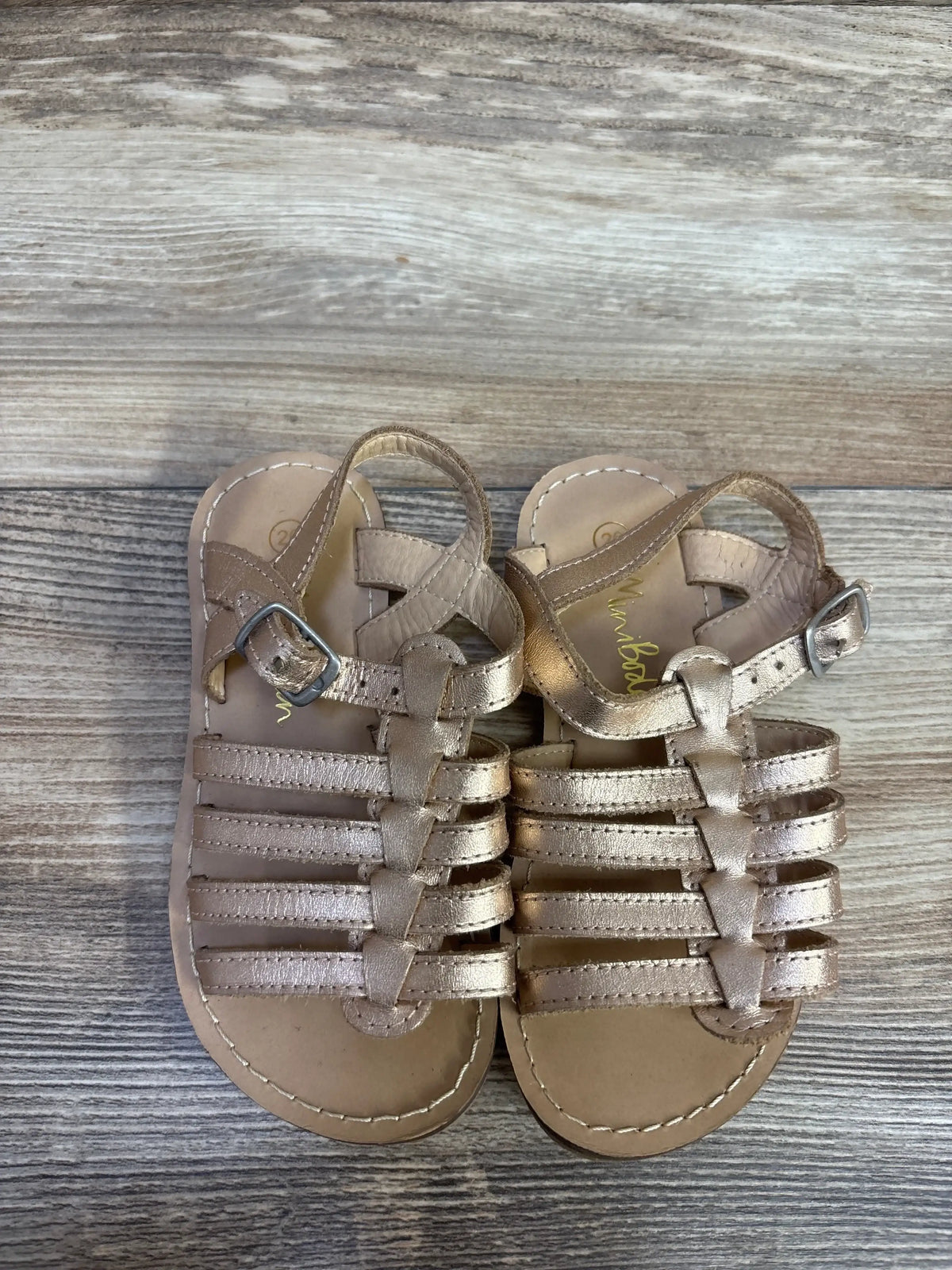 Top down view of Mini Boden gladiator sandals in metallic gold showing strappy front and ankle buckle on wood floor