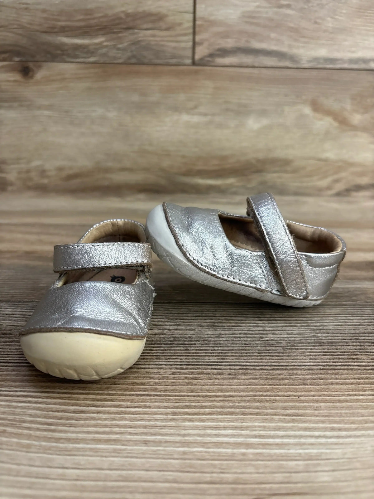 Pair of metallic silver toddler Mary Jane shoes with soft beige soles on wood surface