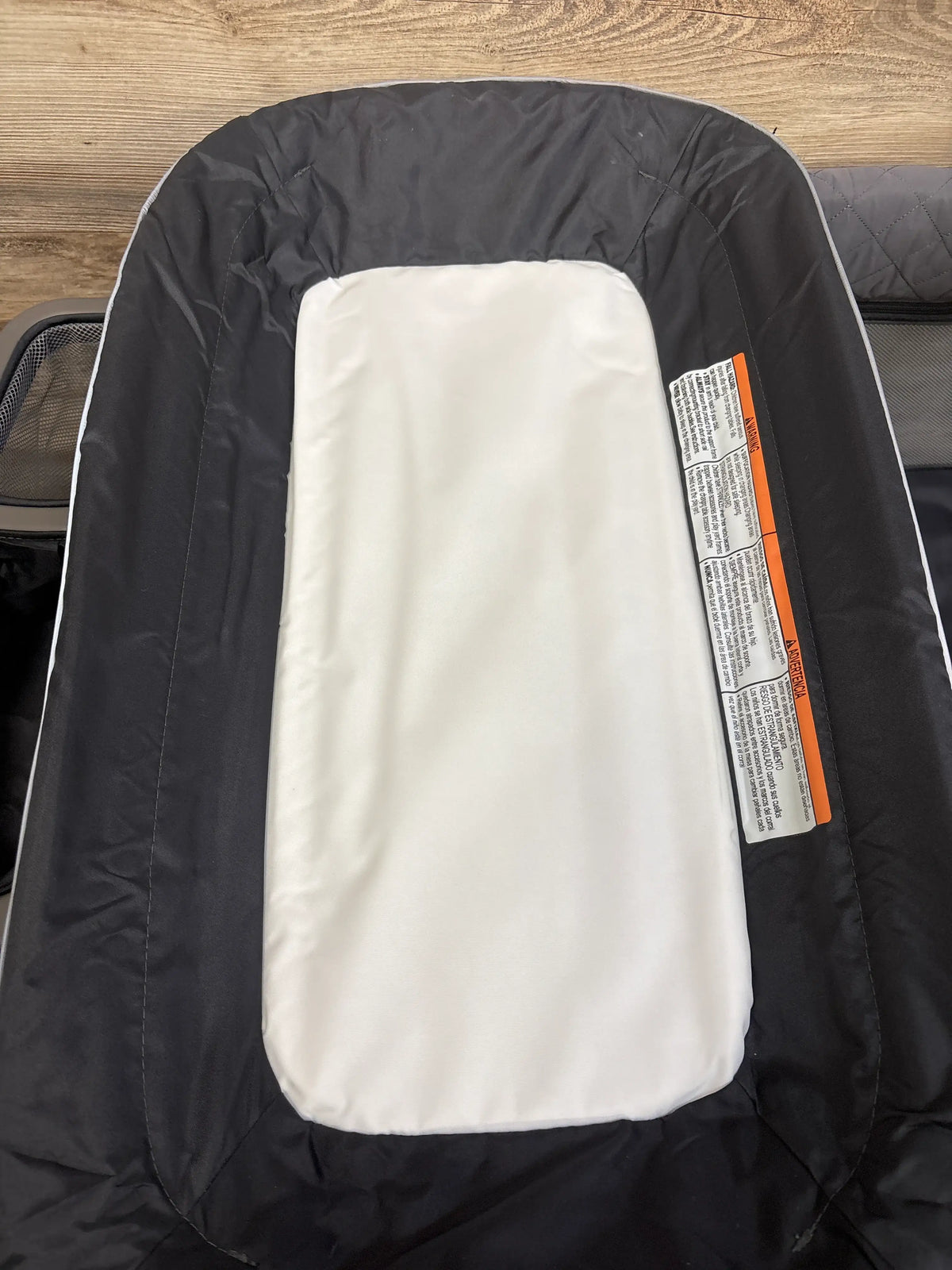 Close-up of the grey Pamo Babe Just Right Playard bassinet showing its white padded insert with a wood floor background