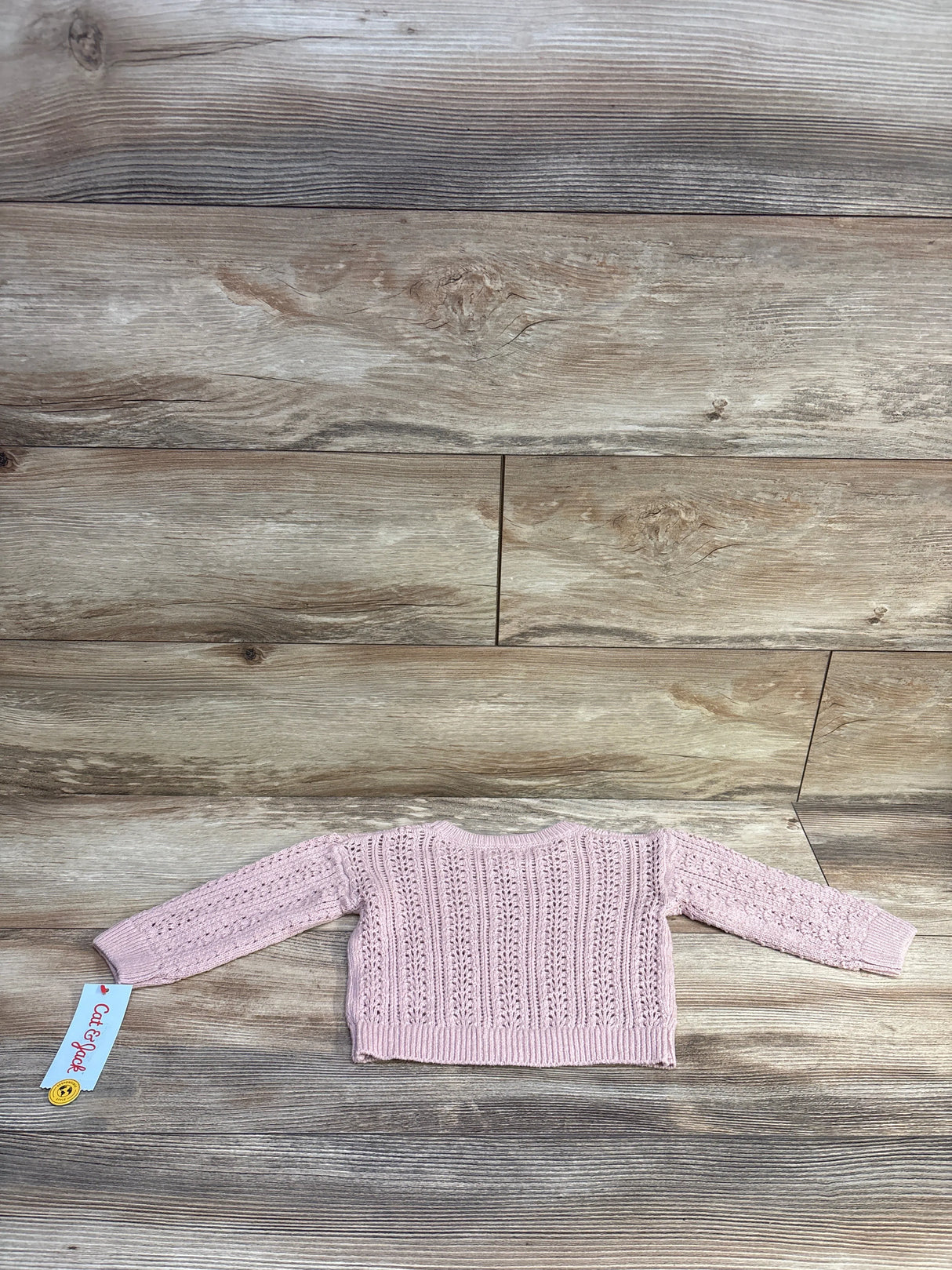 Back view of Cat and Jack pink crochet baby sweater size 12 months showing openwork stitch pattern, ribbed hem and long sleeves on wood background