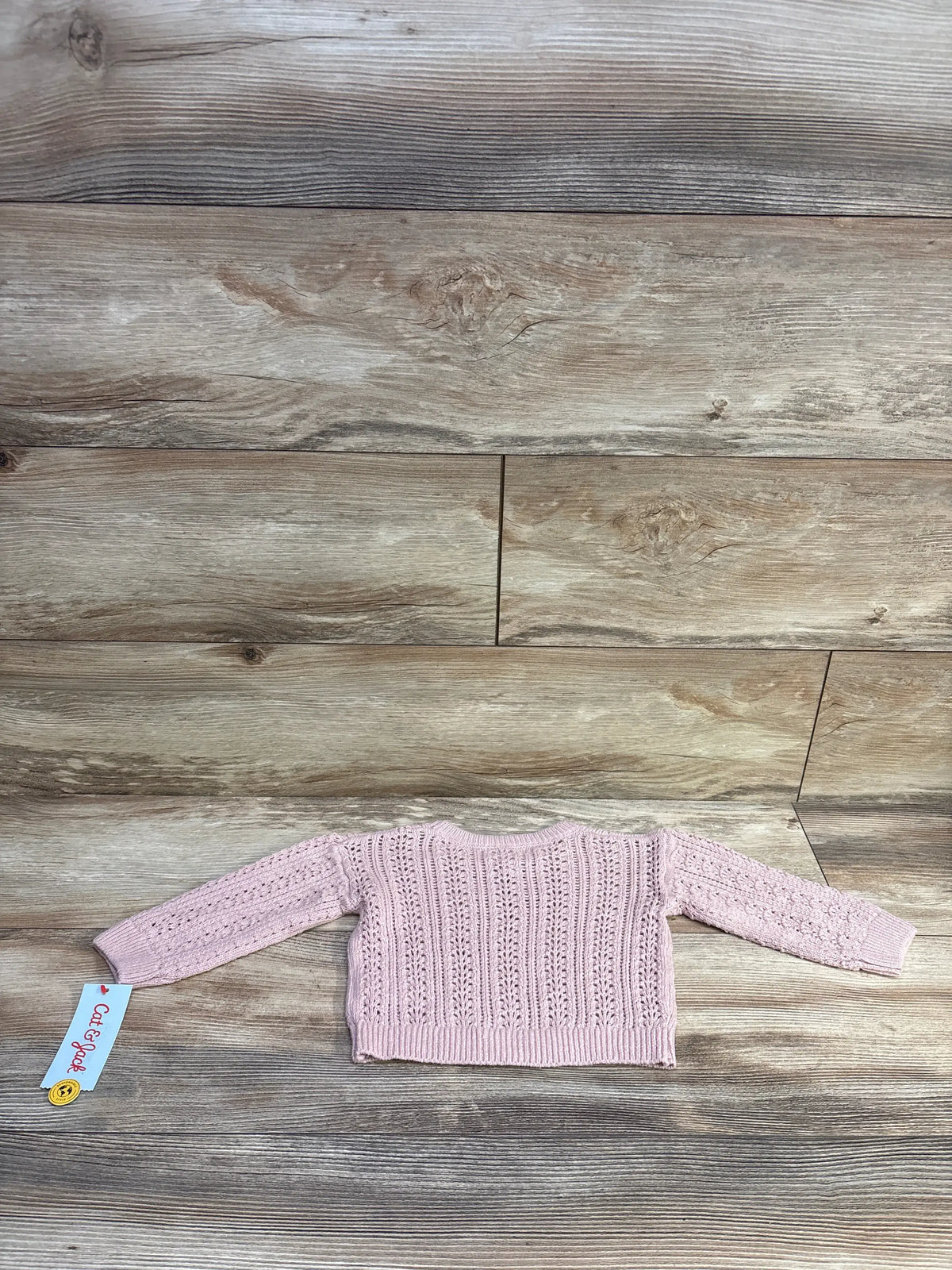 Back view of Cat and Jack pink crochet baby sweater size 12 months showing openwork stitch pattern, ribbed hem and long sleeves on wood background