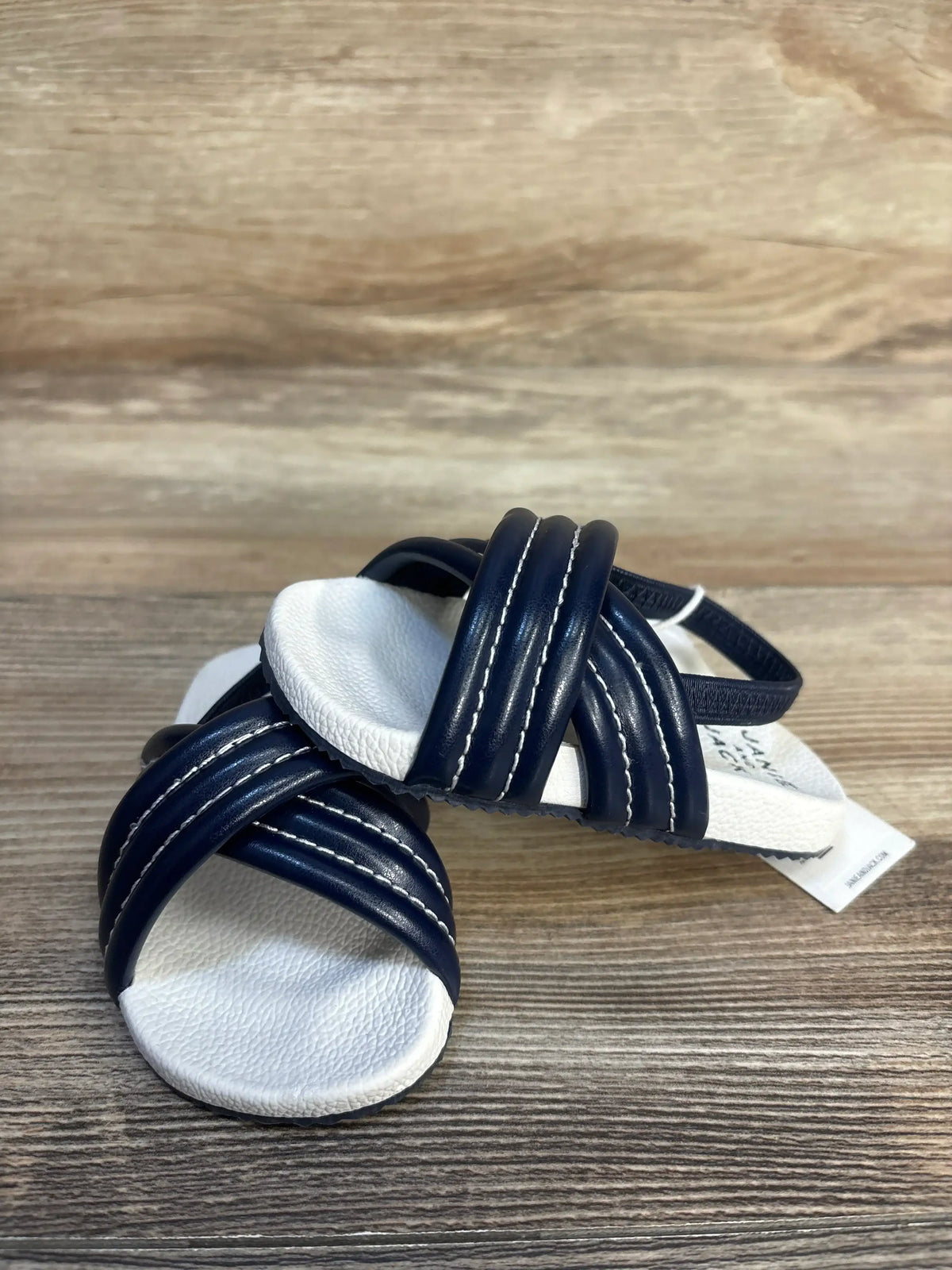 Three-quarter view of Janie and Jack toddler cross-strap pool slide showing navy crisscross straps with white cushioned footbed on wood plank floor