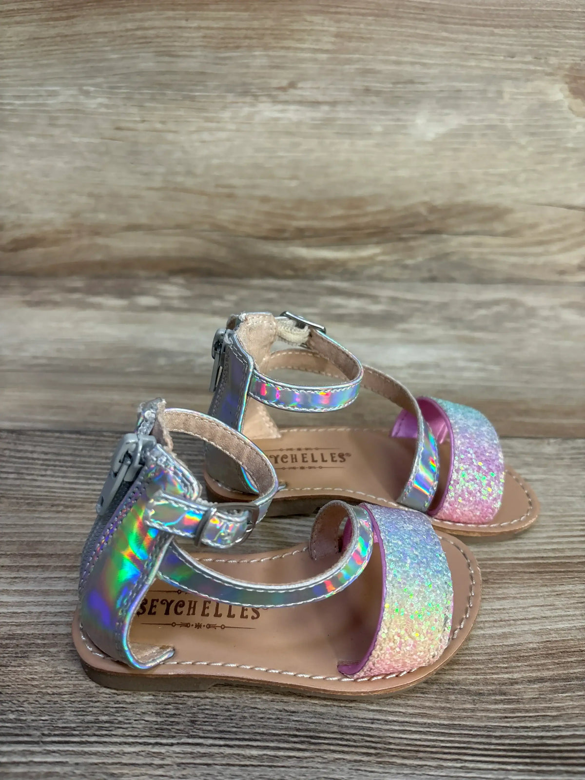 Three-quarter view of Seychelles metallic sandals 5C showing holographic ankle straps and pink glitter toe strap on wood