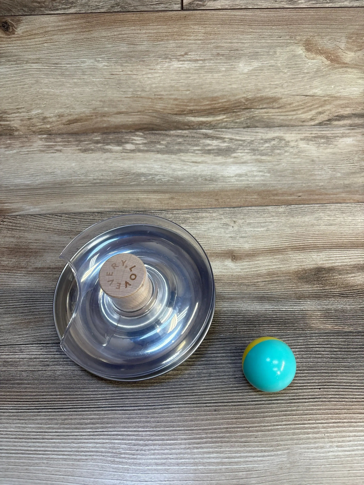 Top-down view of Lovevery Stainless Steel Tip & Turn clear spinner showing wooden knob and teal-and-yellow ball on wood floor
