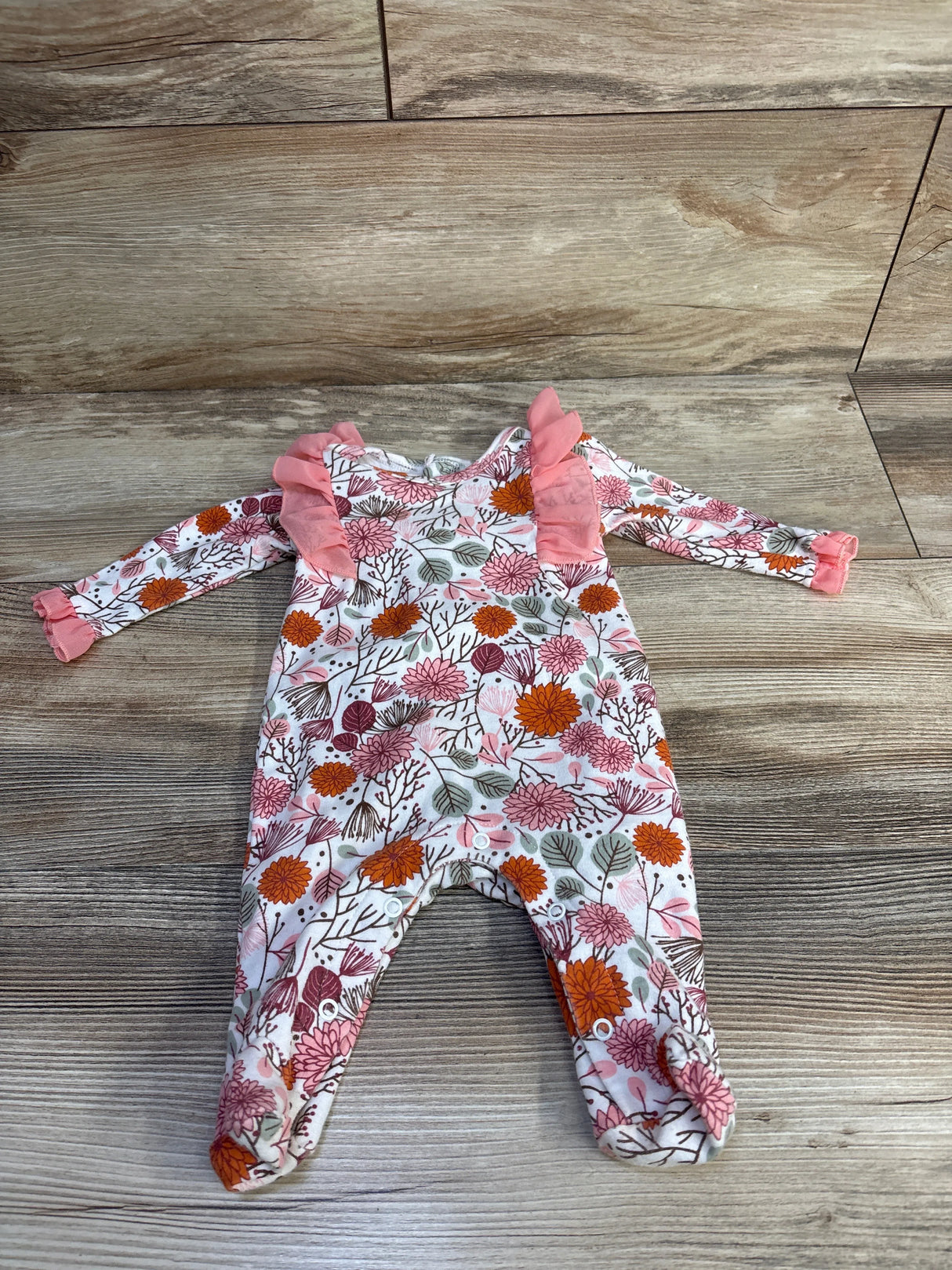 Flat lay of white 3-month baby footed coverall showcasing pink tulle ruffle sleeves and orange-pink floral print on wood background