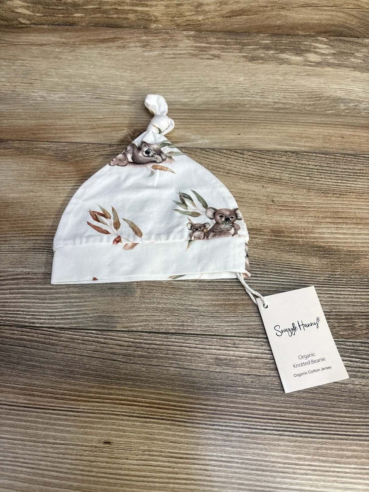 Flat lay of knotted baby beanie showing koala print and Snuggle Hunny tag on wood background, size 0-6 months