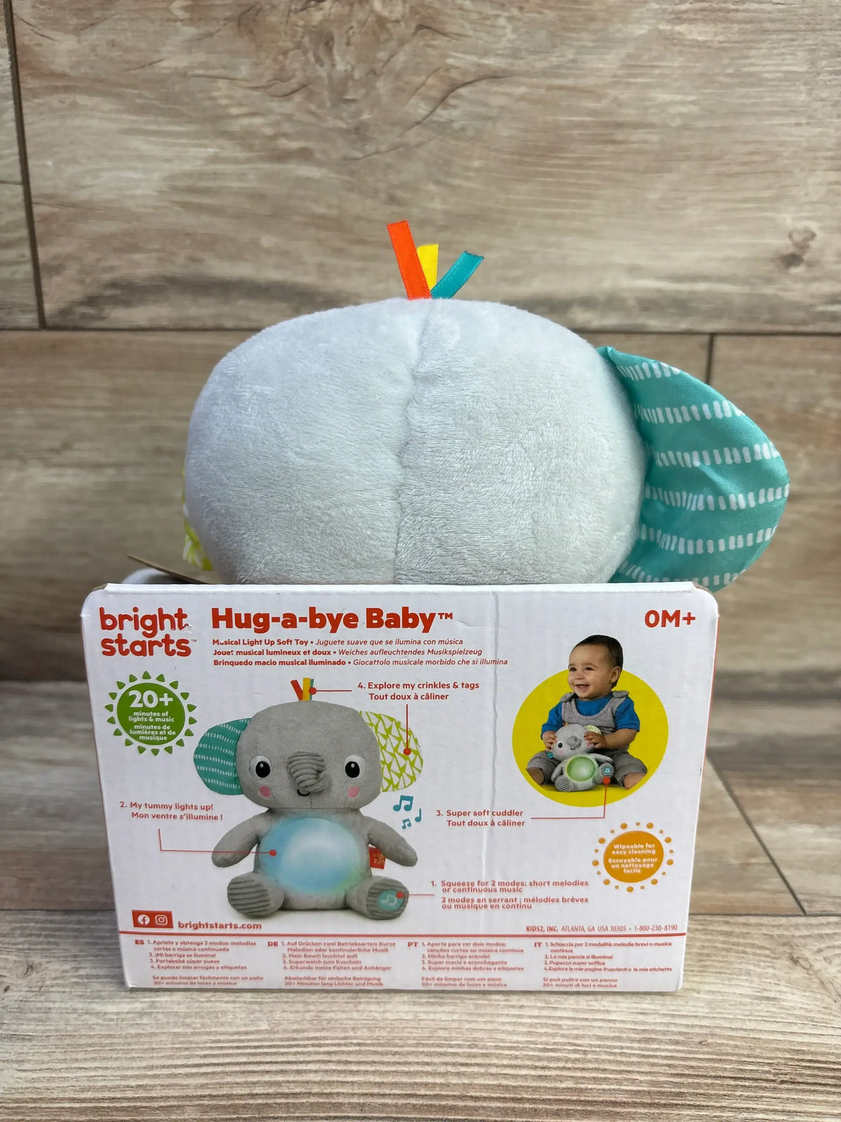Back view of Bright Starts Hug-a-Bye Baby elephant plush with teal ear, ribbon tags, sitting on its box on wood floor