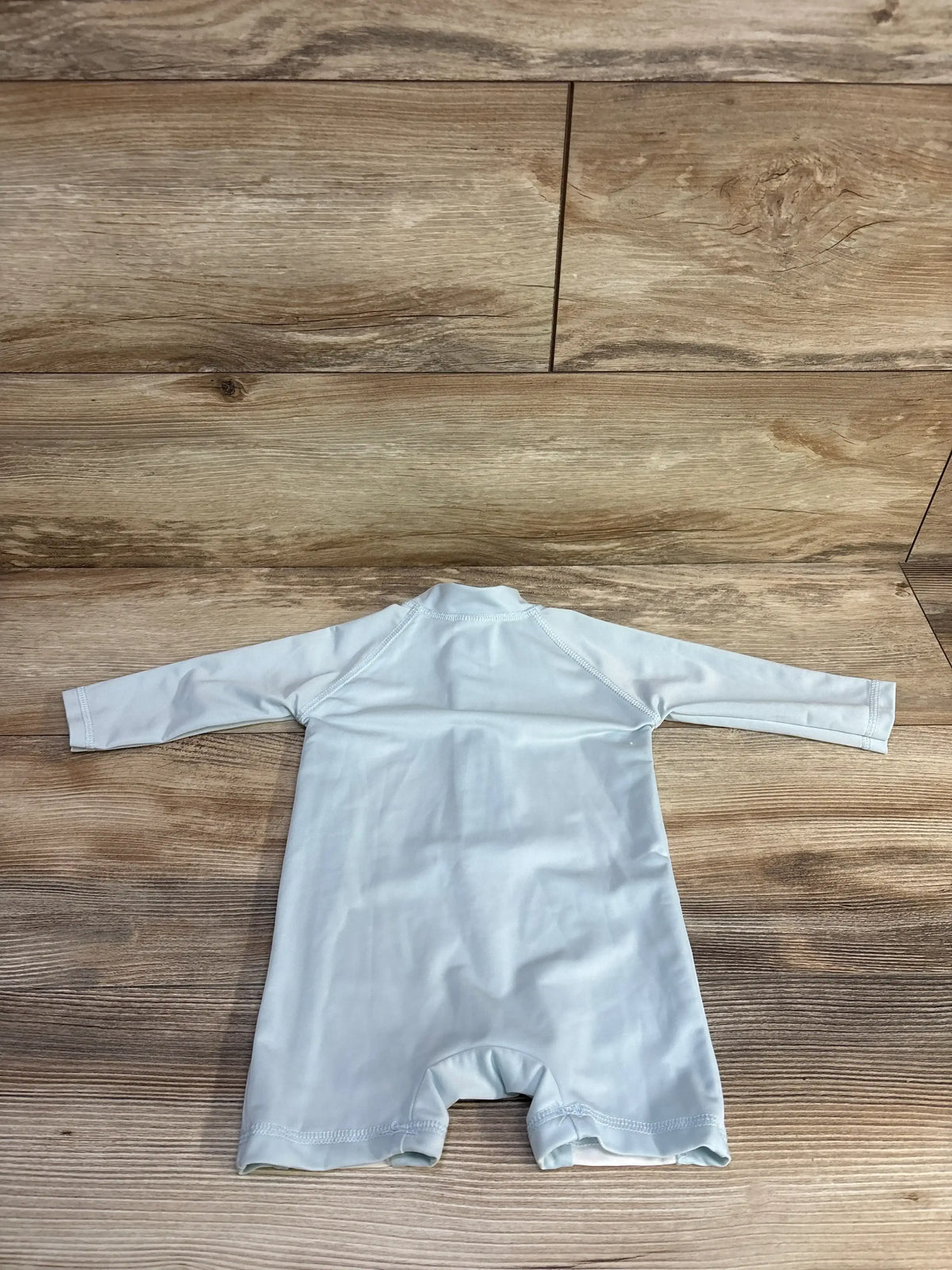 Back view of Cuddle Cubs light blue one-piece rash guard showcasing long sleeves and short legs on a wood-look floor