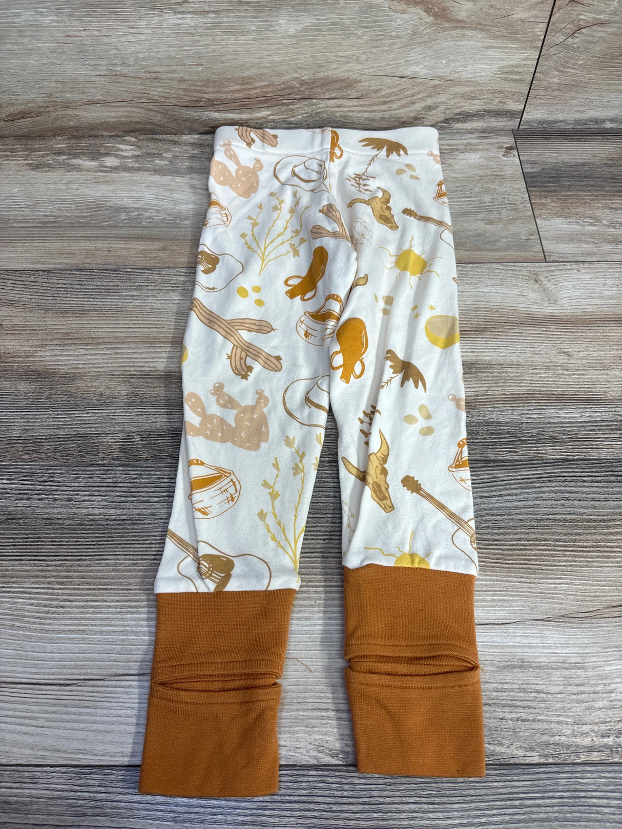 Top-down view of Goumi convertible baby pants (3T) in Western Ivory showing brown fold-over cuffs and western print on wood floor