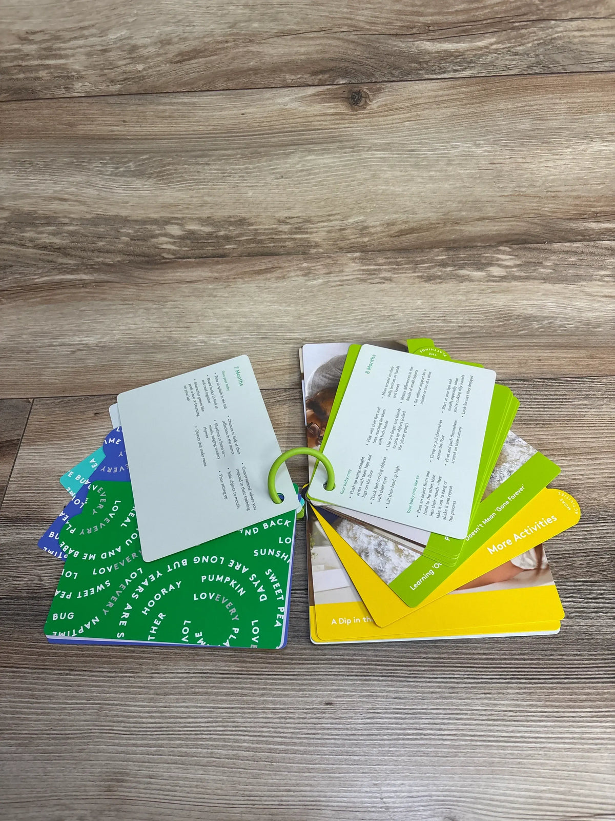 Overhead view of The Lovevery Inspector Play Guide Mint 7-8m fanned to show colorful activity cards on a green ring on a wooden surface