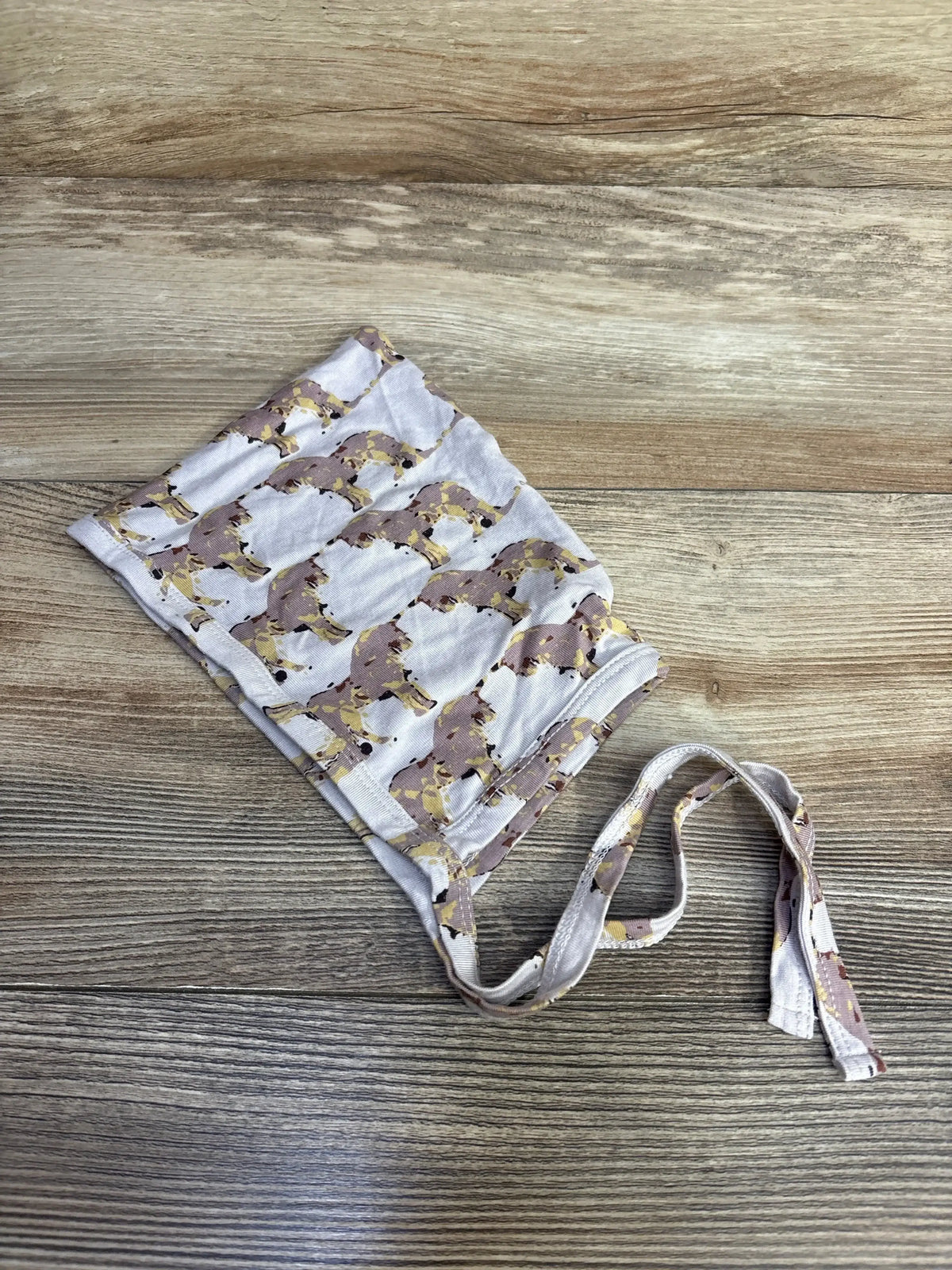 Flat lay of Kate Quinn baby unicorn bonnet size 12–18 months showing pink unicorn print with gold accents and tie straps on wood background