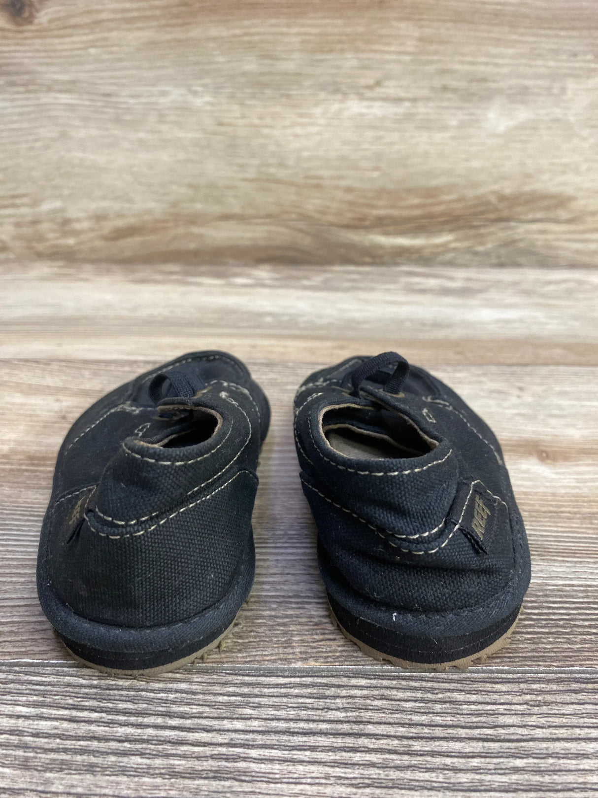 Pair of black kids' canvas slip-on shoes with visible white stitching on wooden background