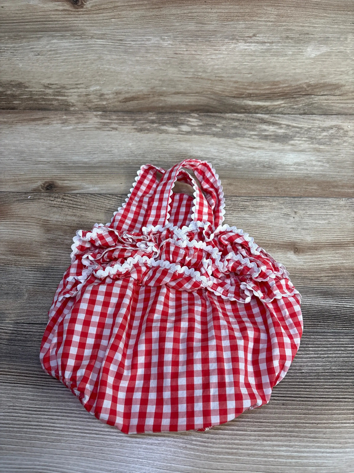 Top down view of red gingham baby romper 12 months with ruffled white trim and straps on wood background