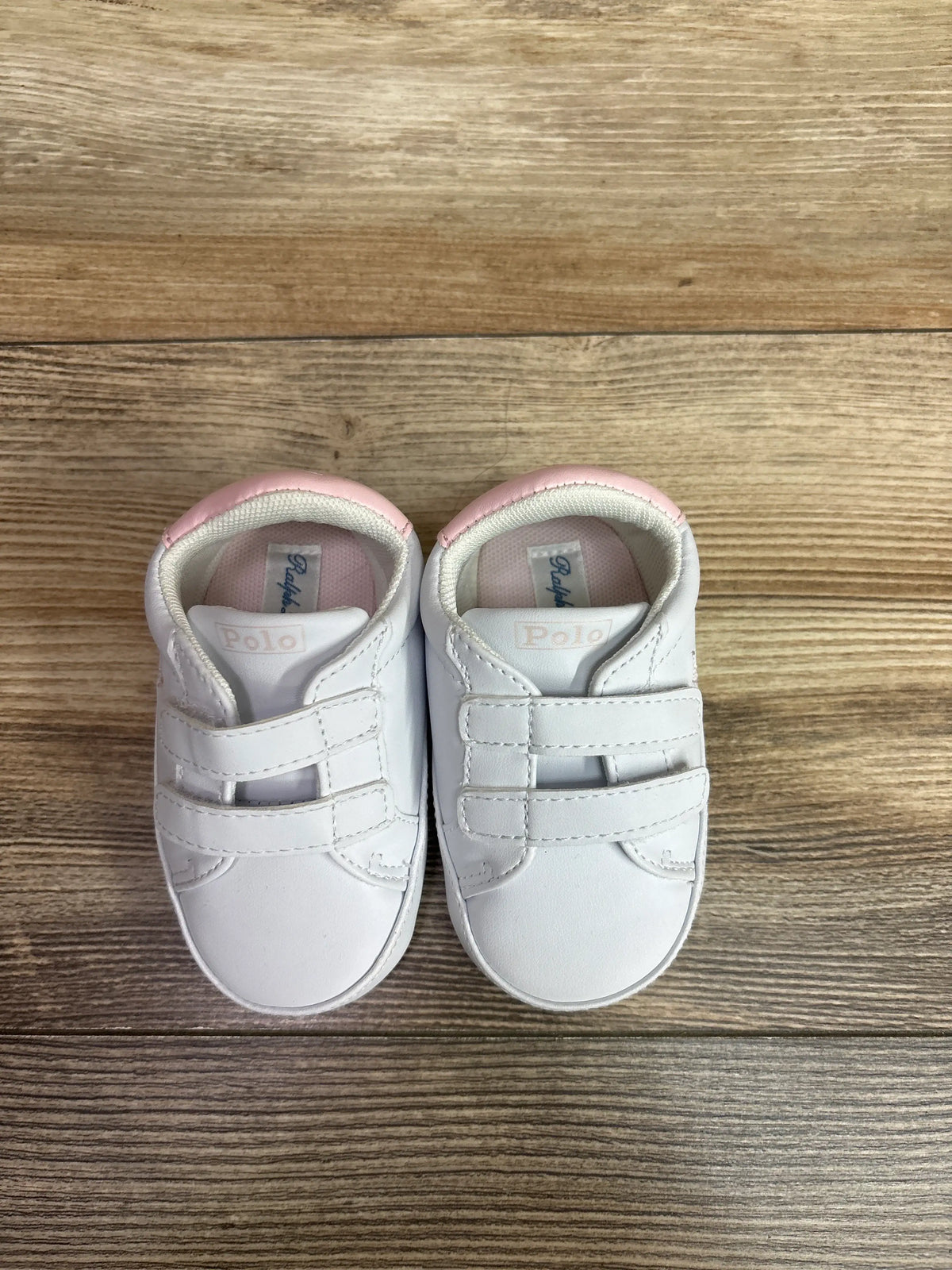 Top view of white Polo Ralph Lauren Heritage Court II EZ crib sneakers showcasing dual hook-and-loop straps and pink lining on a wood surface