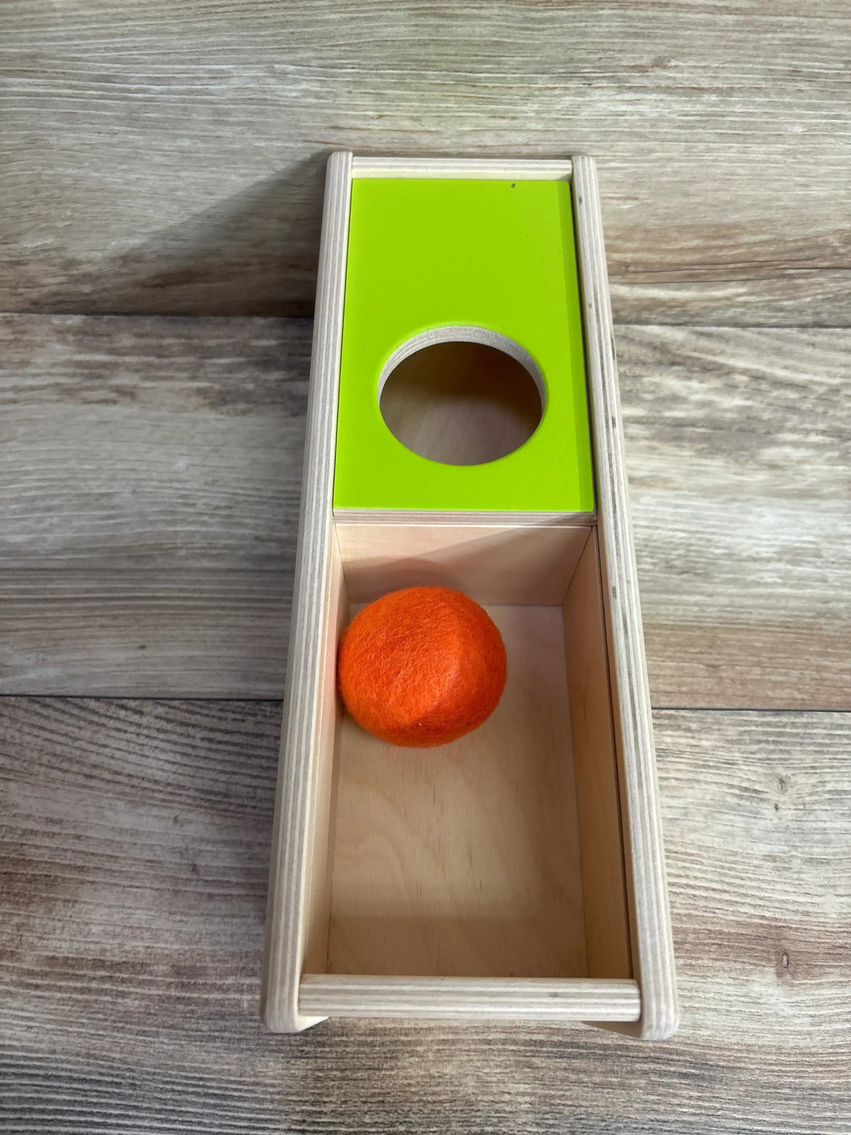 Top-down view of Lovevery Sliding Top Box showing green sliding top with round hole and orange felt ball inside wooden box on wood surface